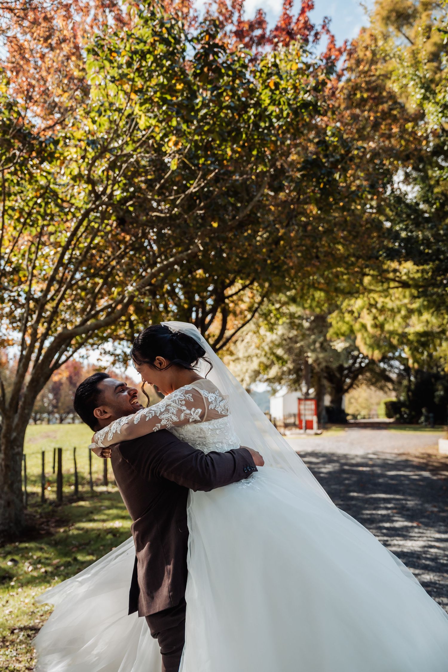 Groom lifting bride in a white lace gown for a kiss outdoors among colorful autumn trees.