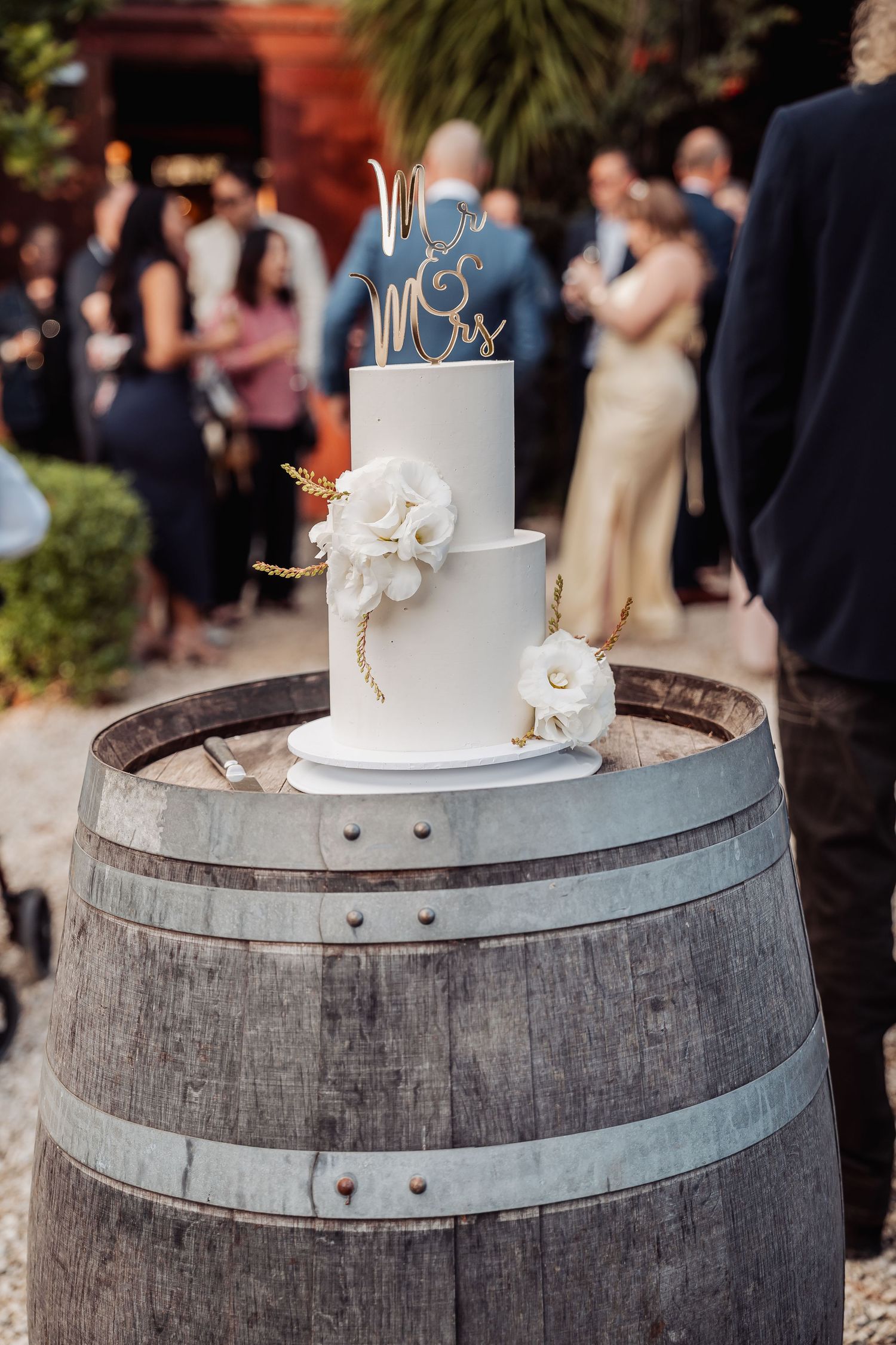 Elegant white two-tier wedding cake with silver Mr & Mrs topper displayed on rustic wine barrel at outdoor reception.