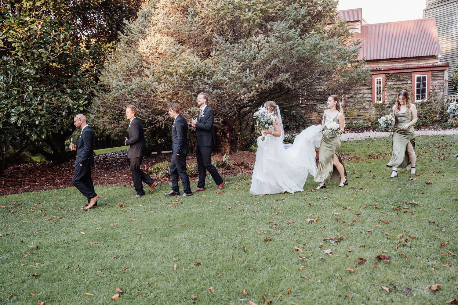Wedding party walking and dancing on a grass lawn outside a rustic brick building during an outdoor celebration.