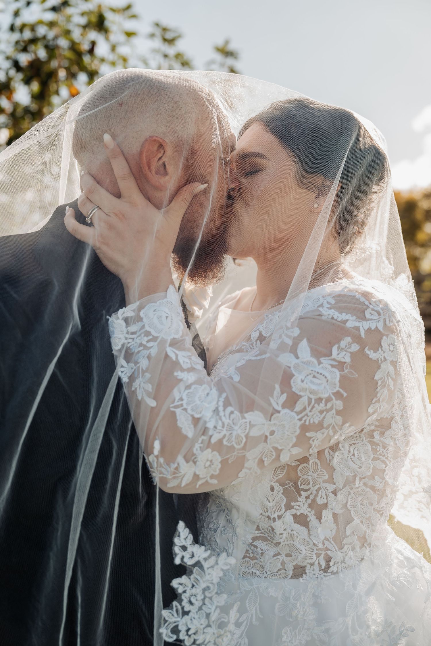 Intimate wedding portrait captures couple sharing kiss under delicate lace veil in natural light.