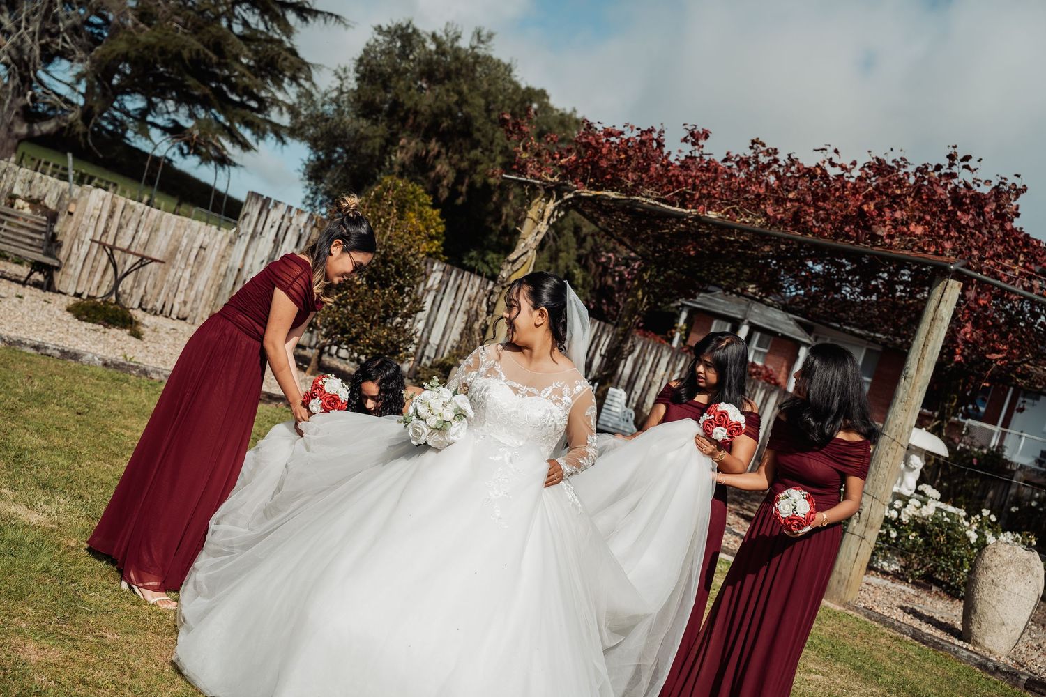 Bride in white gown with bridesmaids in burgundy dresses arranging her dress outdoors at wedding.
