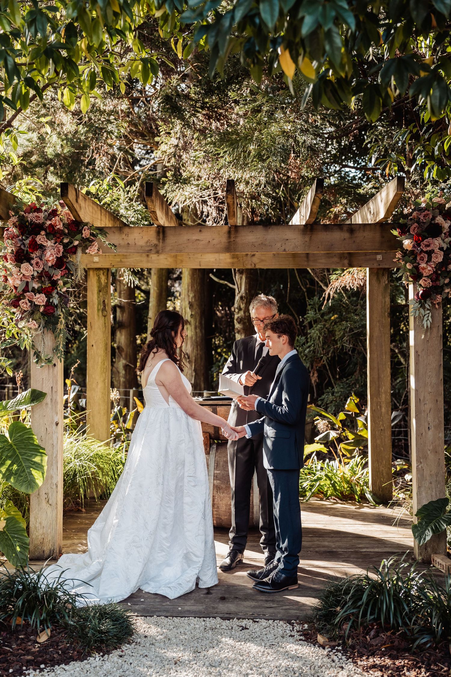 Bride and groom exchange vows under a rustic wooden pergola adorned with pink florals in a lush garden ceremony.