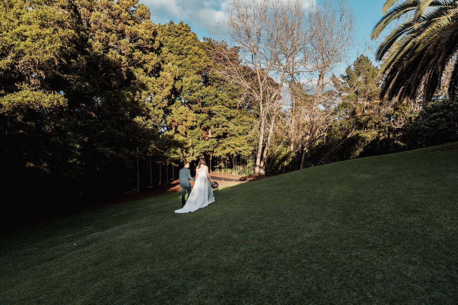 Bride and groom walk across sunlit green lawn surrounded by tall trees at wedding.