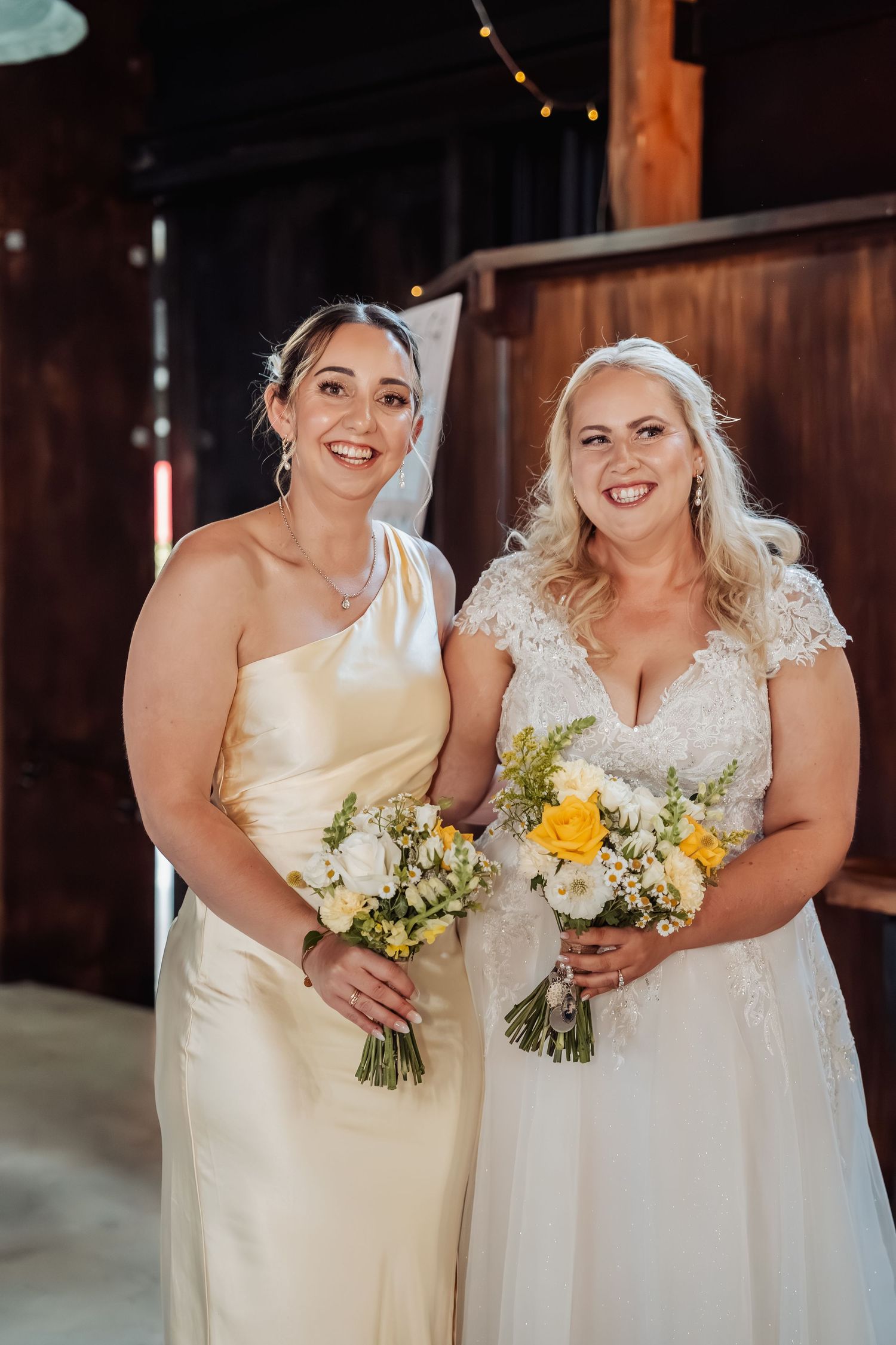 Two brides smile together in a rustic barn, one in a lace gown and one in a satin dress, holding bouquets.
