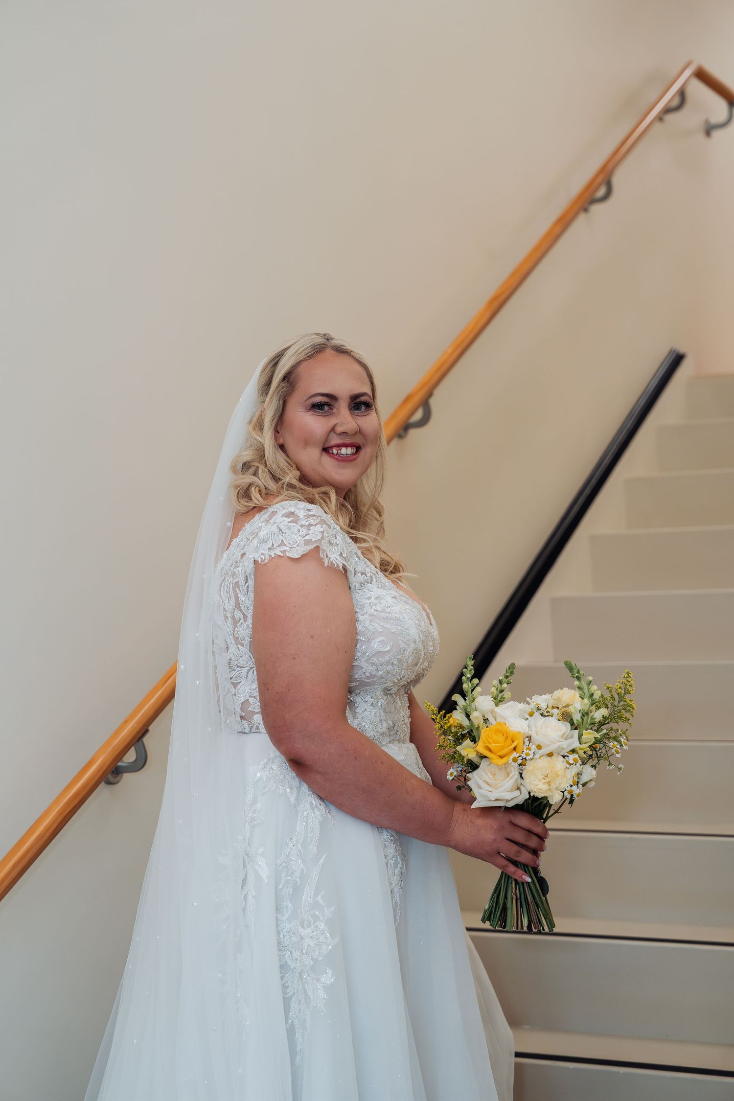 Blonde bride in lace wedding gown holding yellow and white bouquet on staircase.