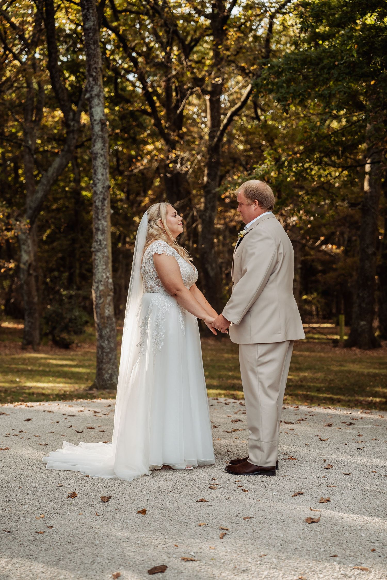 Bride in white lace gown and groom in tan suit hold hands on gravel path surrounded by autumn trees.