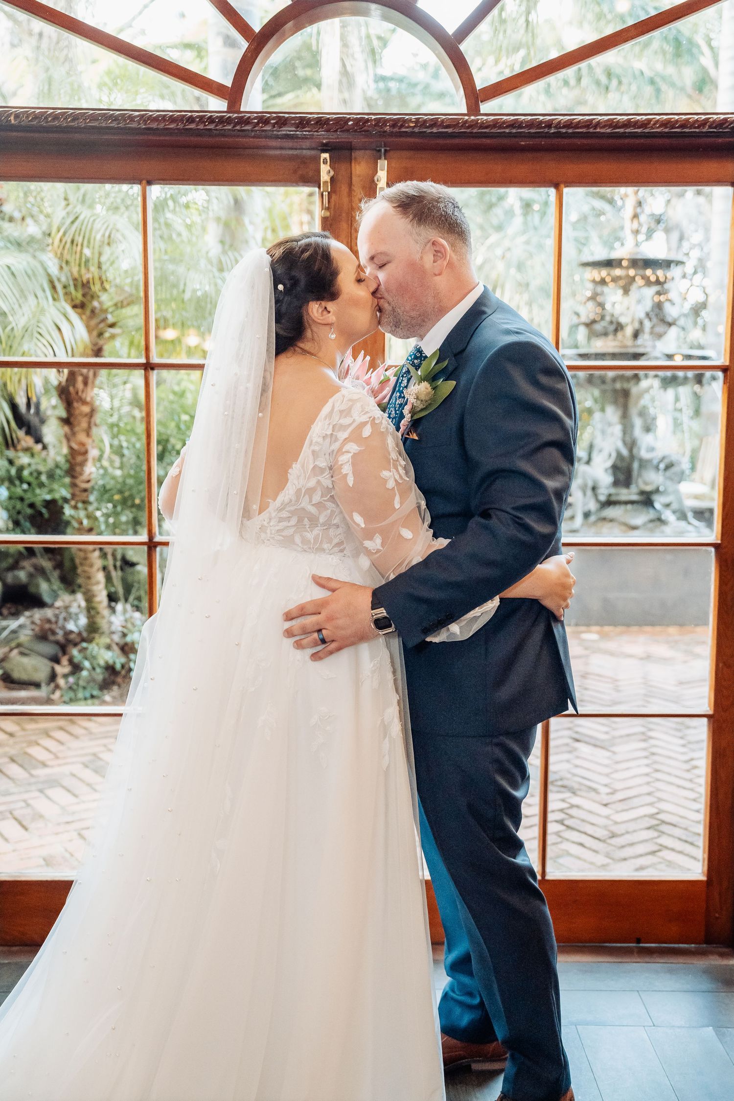 Romantic wedding couple shares intimate moments in a sunlit conservatory with wooden beams and glass panels.