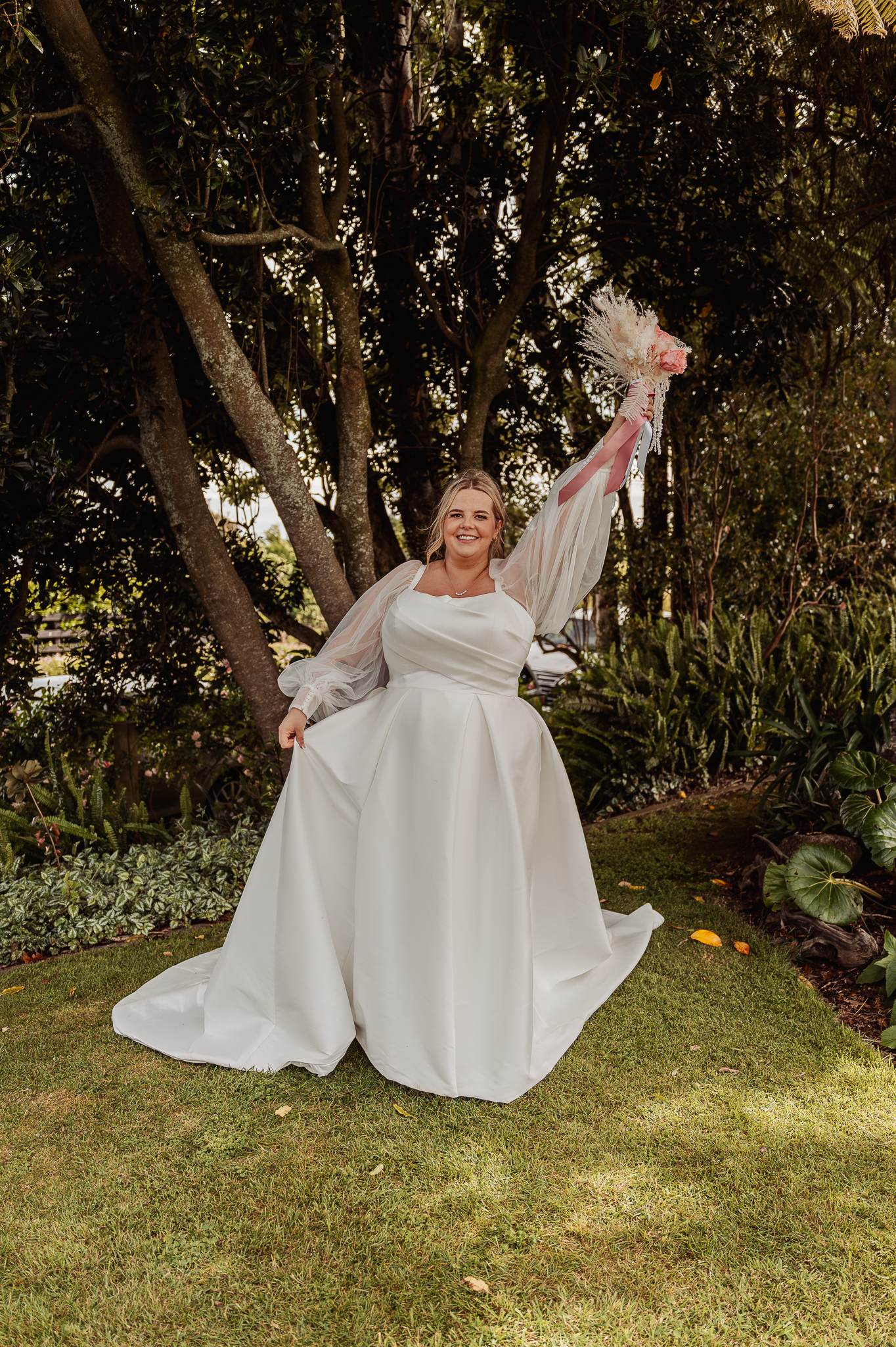 Joyful bride twirling in a flowing white wedding dress on a manicured lawn surrounded by trees.