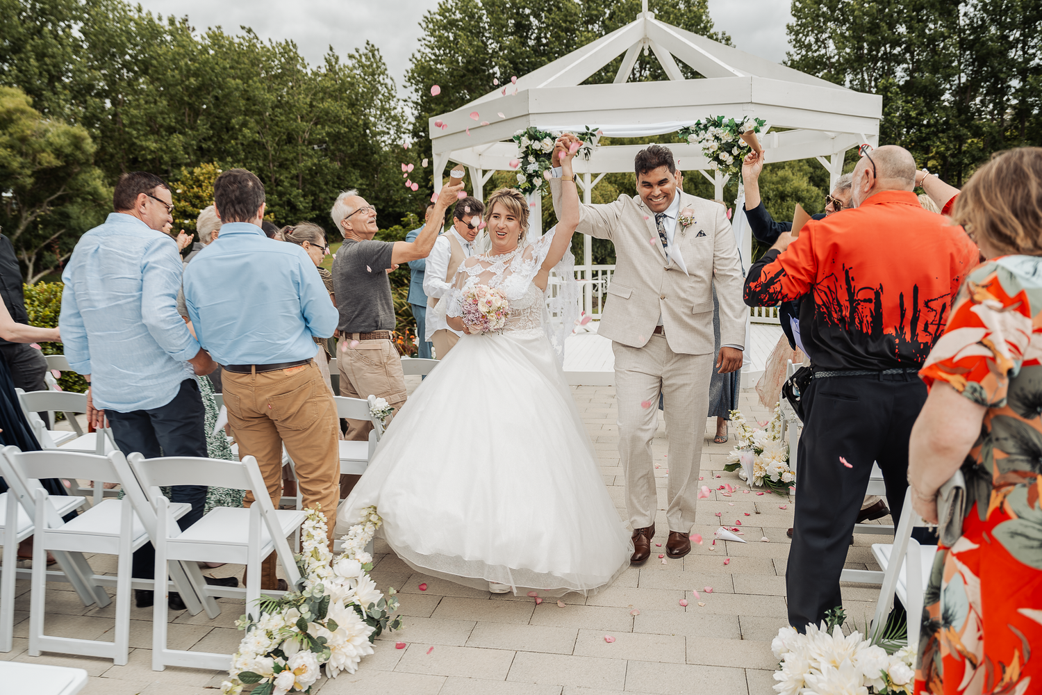 Newlyweds exit their ceremony under a decorated white gazebo while guests celebrate.