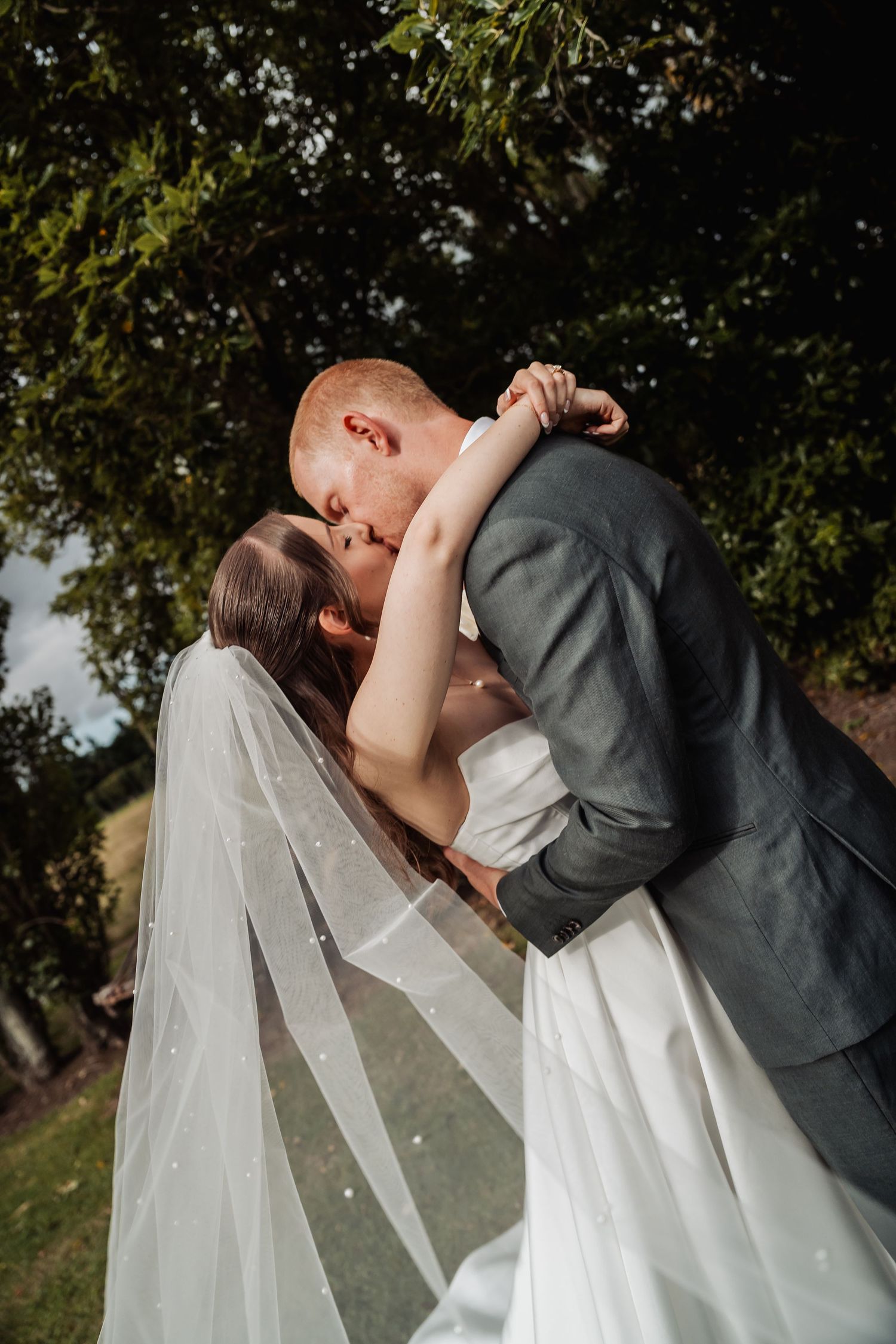 Groom dips bride for a romantic kiss outdoors, her long veil cascading down beneath lush green trees.