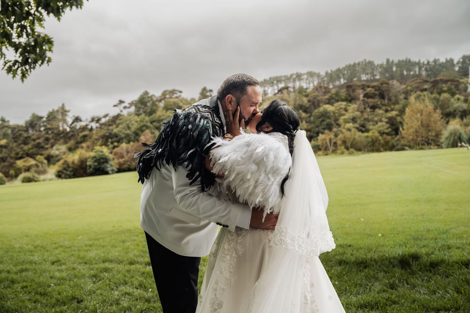 Newlyweds share a kiss in lush green field, groom in white jacket with black feathers, bride in feathered wrap.