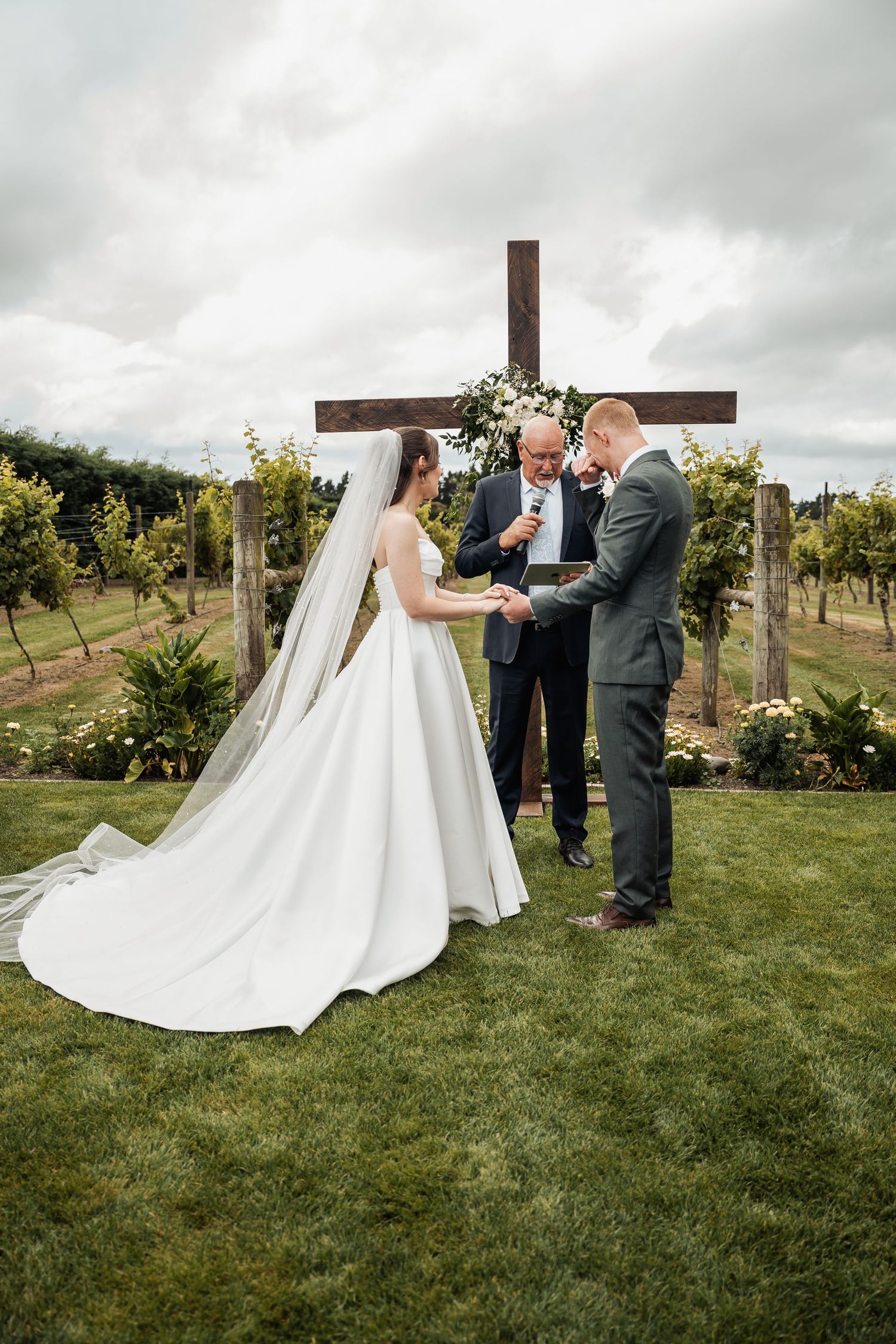 Bride and groom exchange vows before an officiant at an outdoor vineyard wedding ceremony beneath a wooden cross.