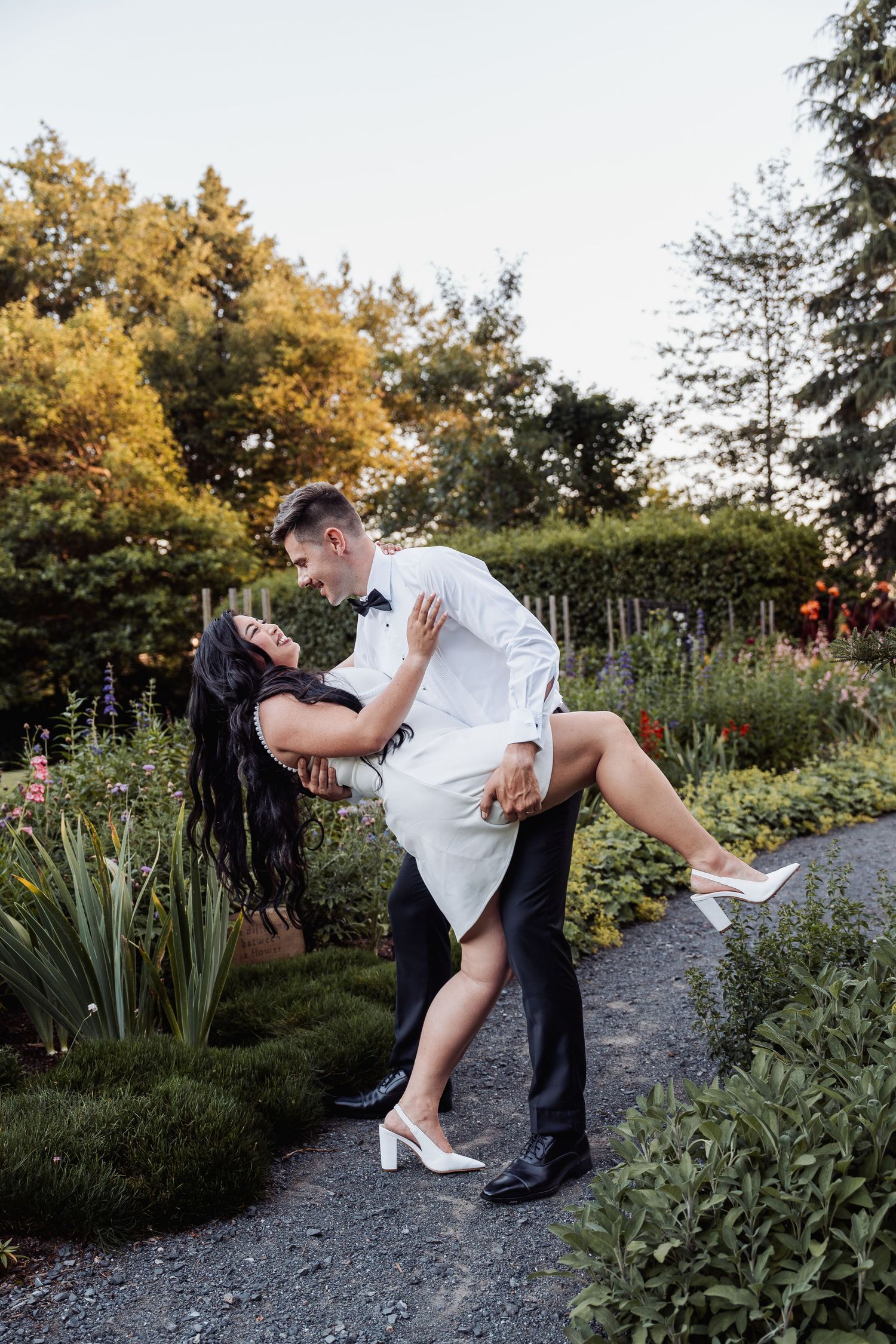 Groom in black tuxedo dips bride in short white dress along garden path at sunset.