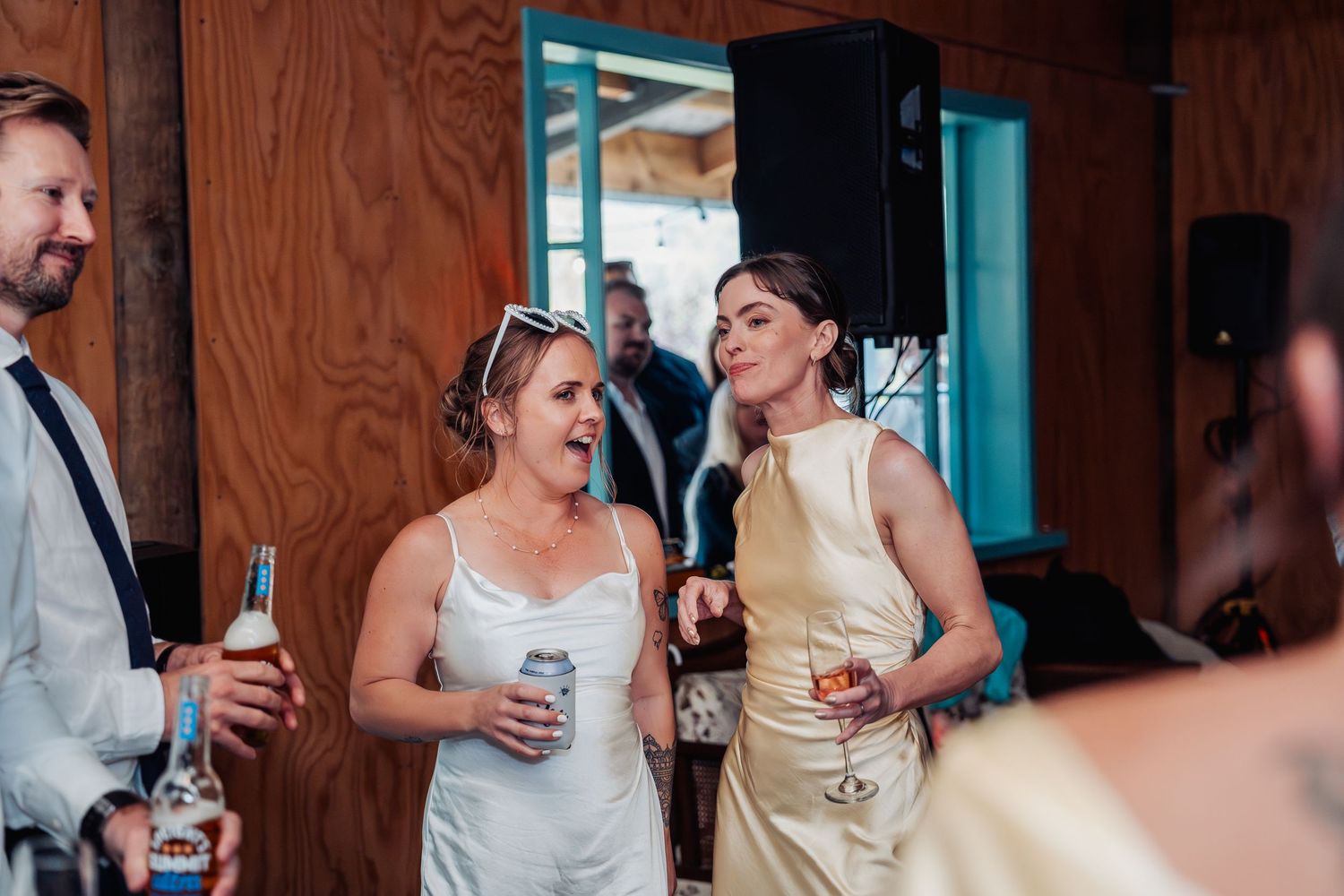 Two women in elegant dresses laughing and holding drinks at a wedding reception.