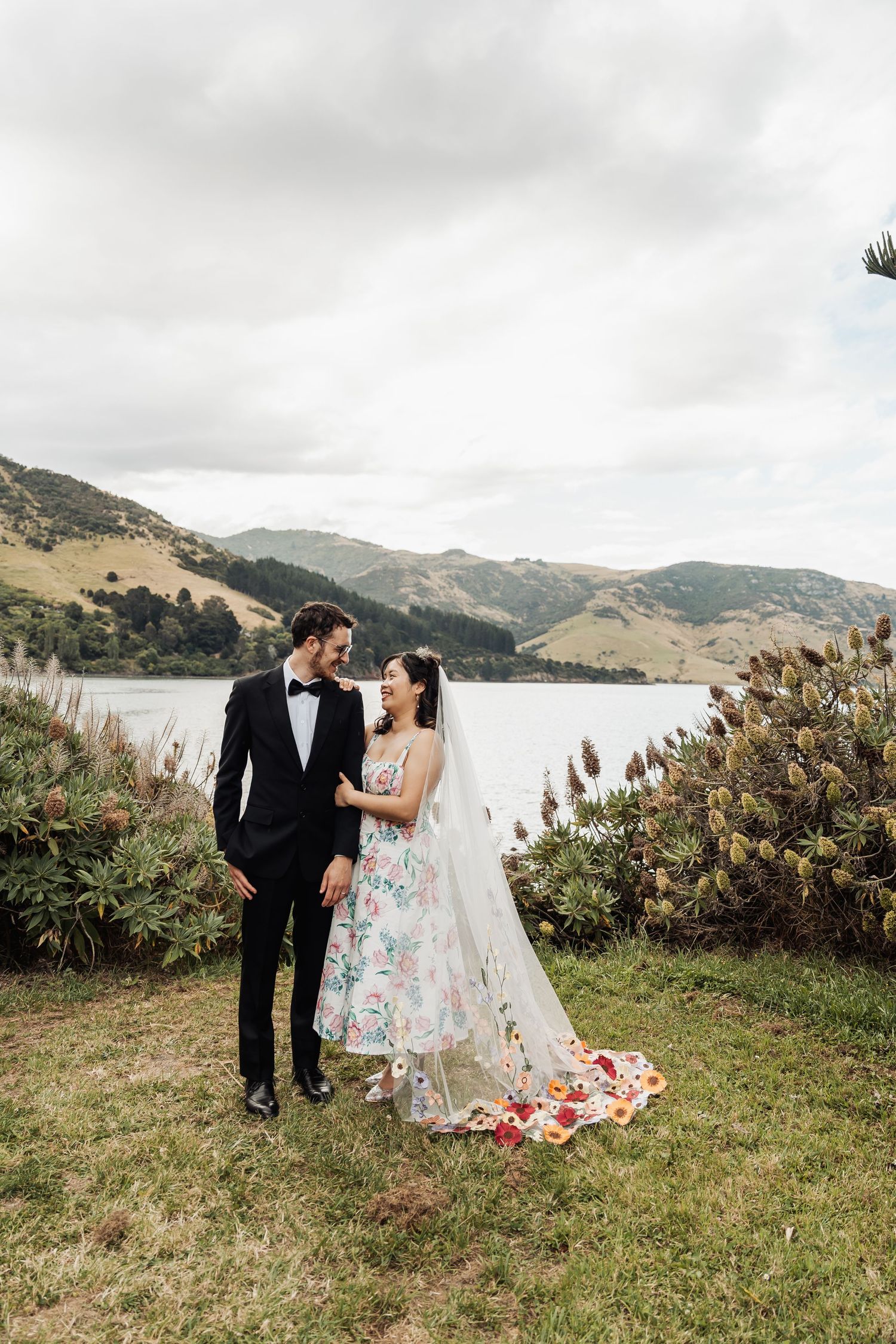 Bride in a floral wedding dress and groom in black tuxedo standing by a serene lake with mountain views.