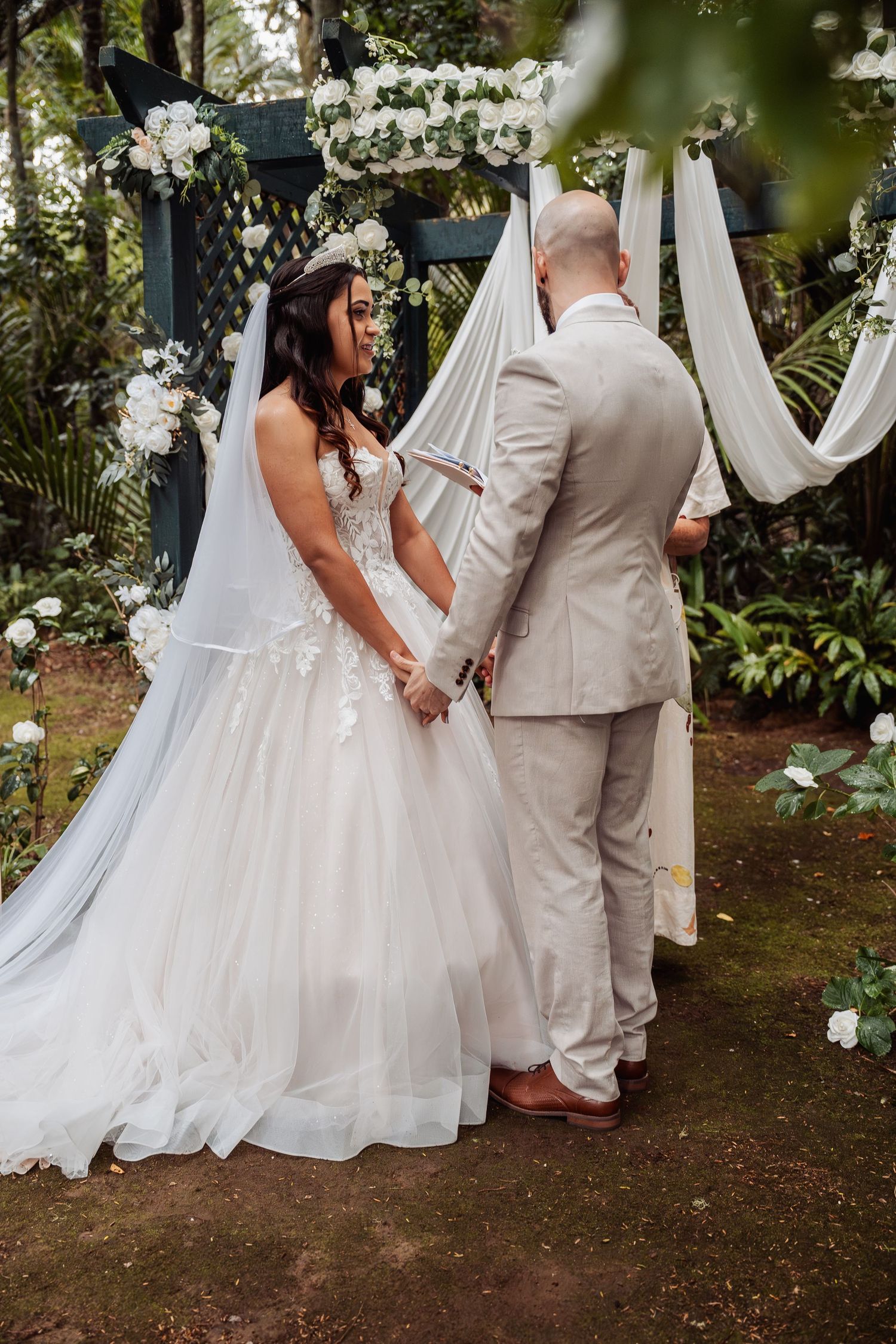 Bride in ball gown and groom in beige suit exchange vows under floral arch in lush garden setting.