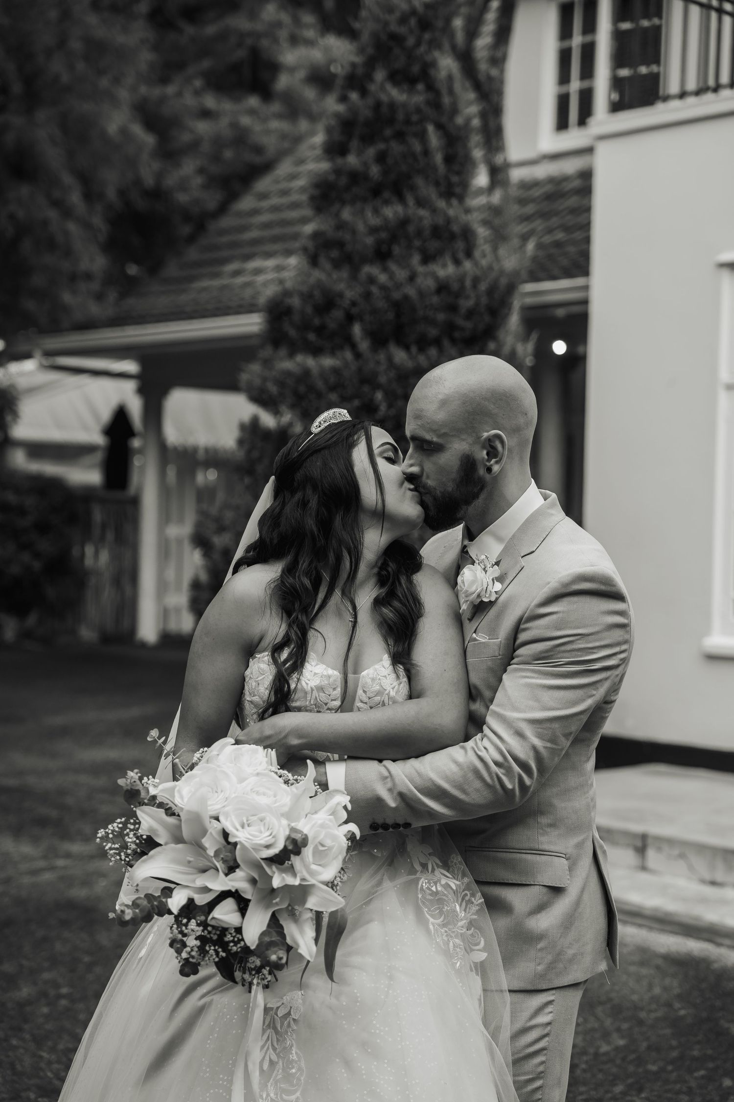 Black and white portrait of bride and groom kissing outdoors with bouquet of white roses.