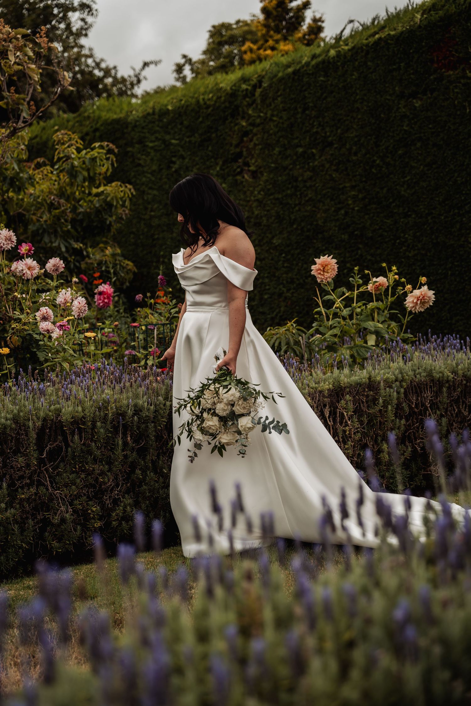 Bride in elegant off-shoulder white gown walks through a lush garden with white bouquet.