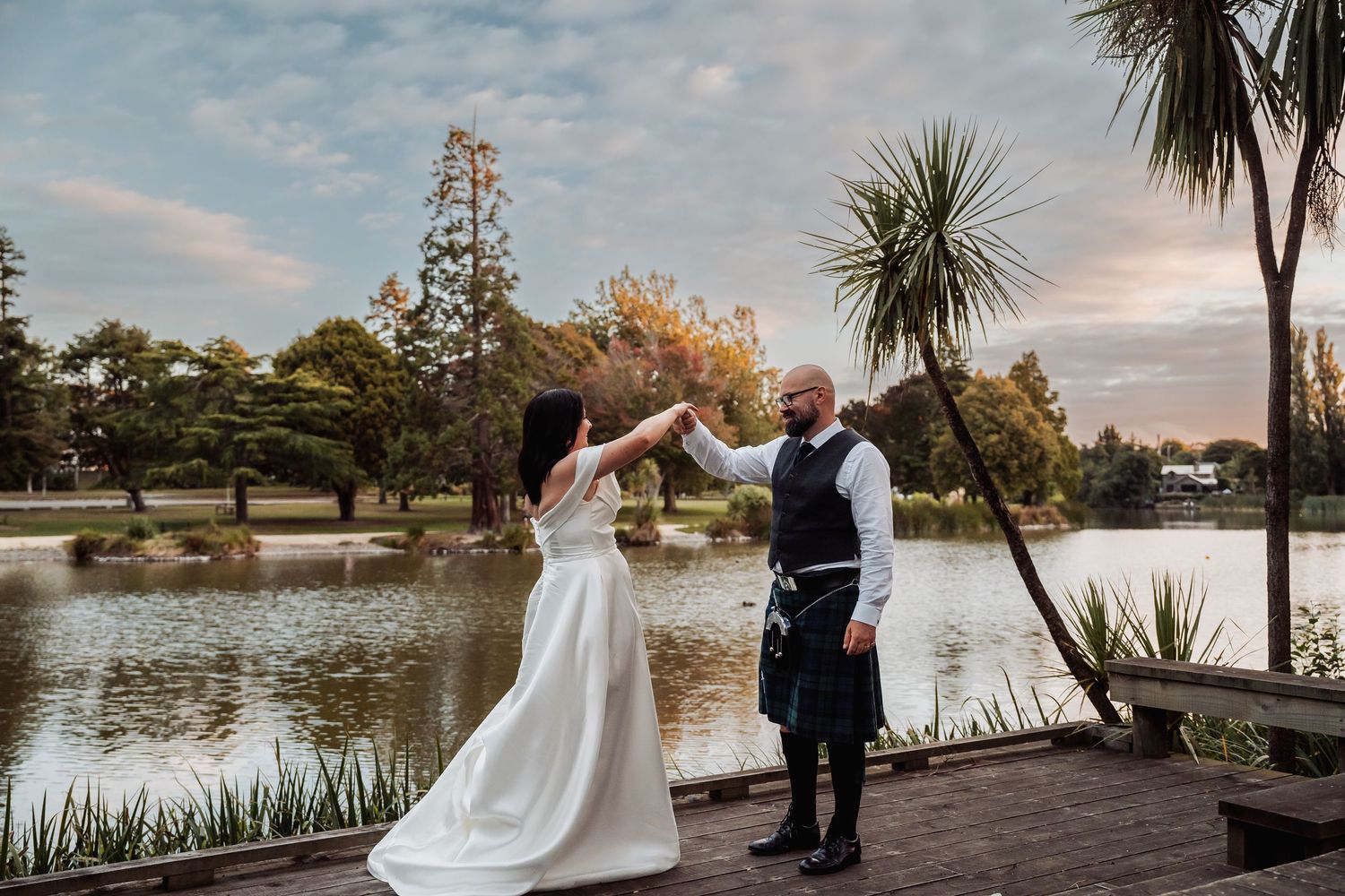 A bride and groom in a kilt dance together on a wooden deck beside a lake at sunset.