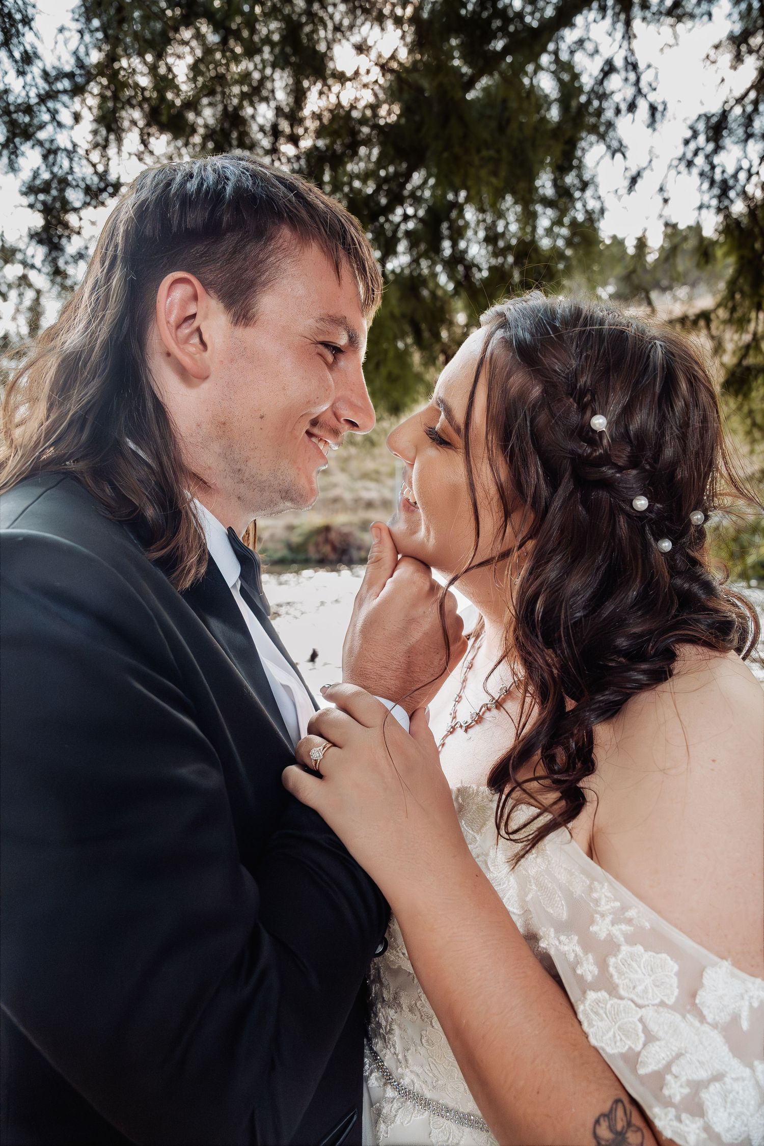 Bride and groom share a romantic close moment, nearly kissing under trees in soft natural light on their wedding day.