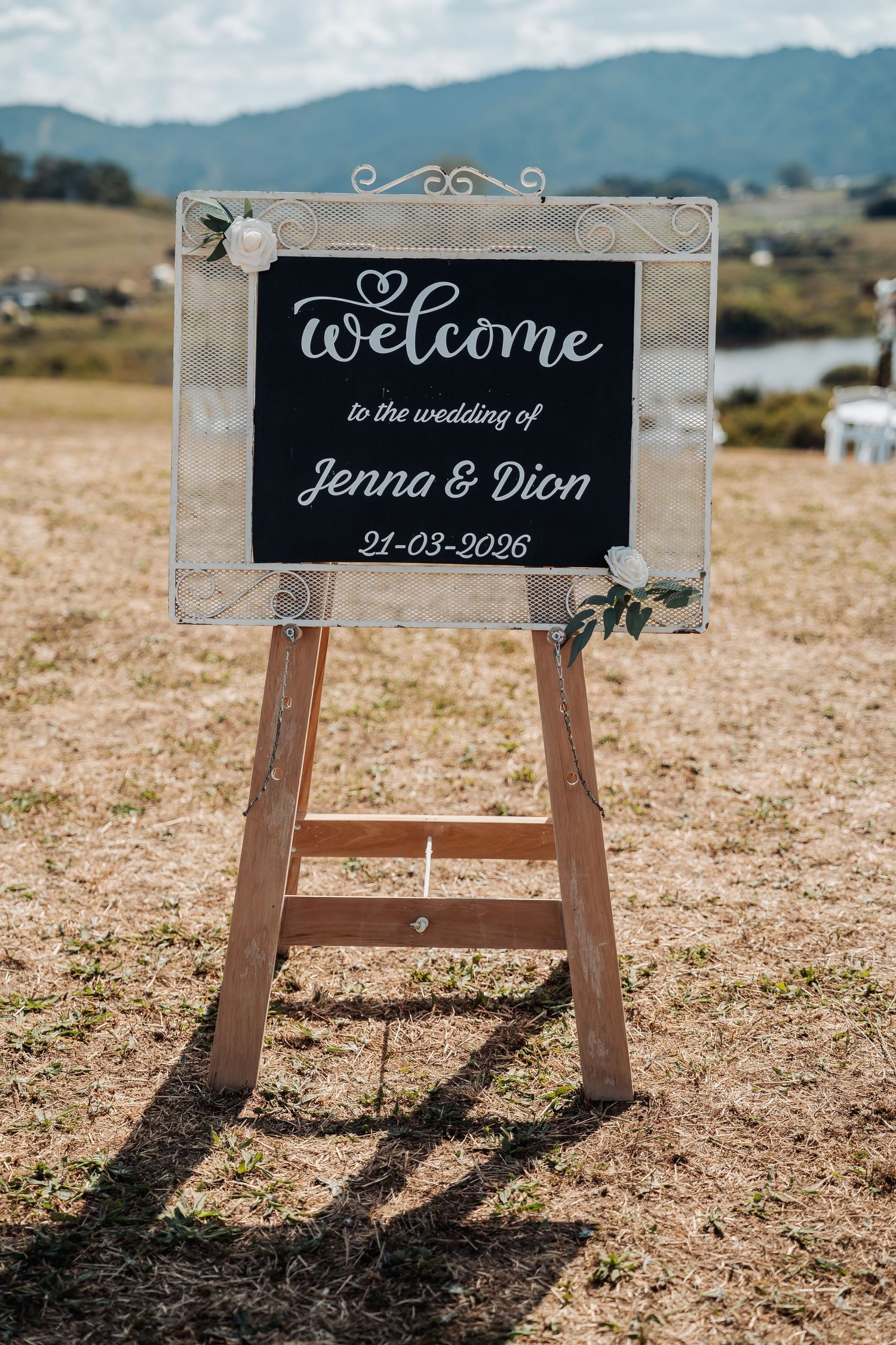 Rustic chalkboard welcome sign on wooden easel reads Welcome to the wedding of Jenna and Dion 21-03-2026.