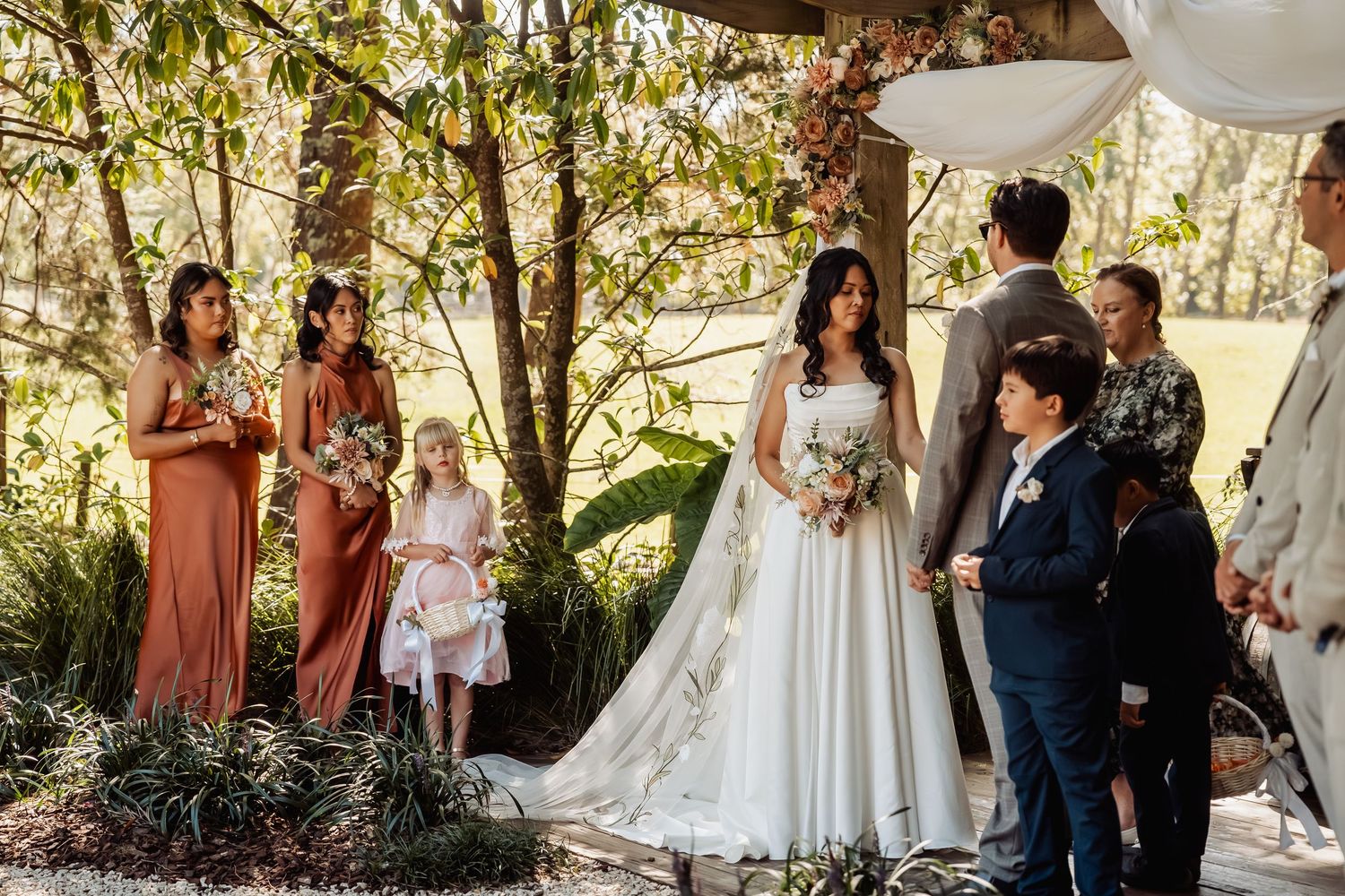 Bride and groom stand at outdoor woodland ceremony altar with bridesmaids in rust dresses nearby.