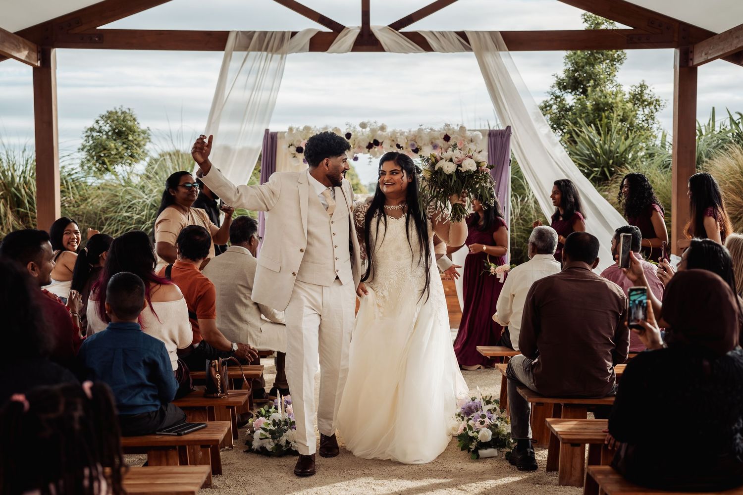 Joyful newlyweds walking down the aisle under a decorated gazebo, celebrated by seated wedding guests.
