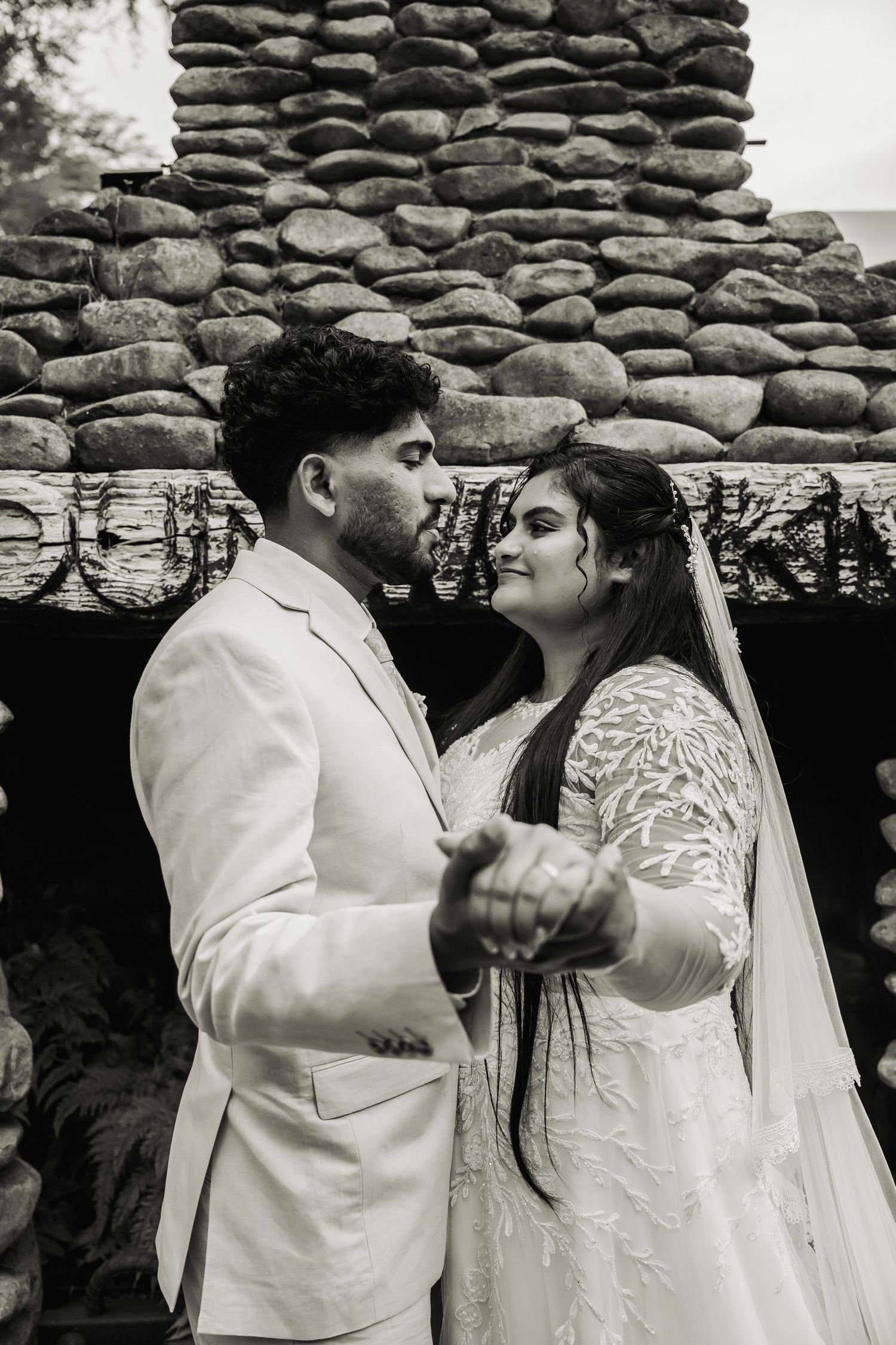Bride and groom hold hands and share a tender moment in front of a rustic stone wall backdrop.