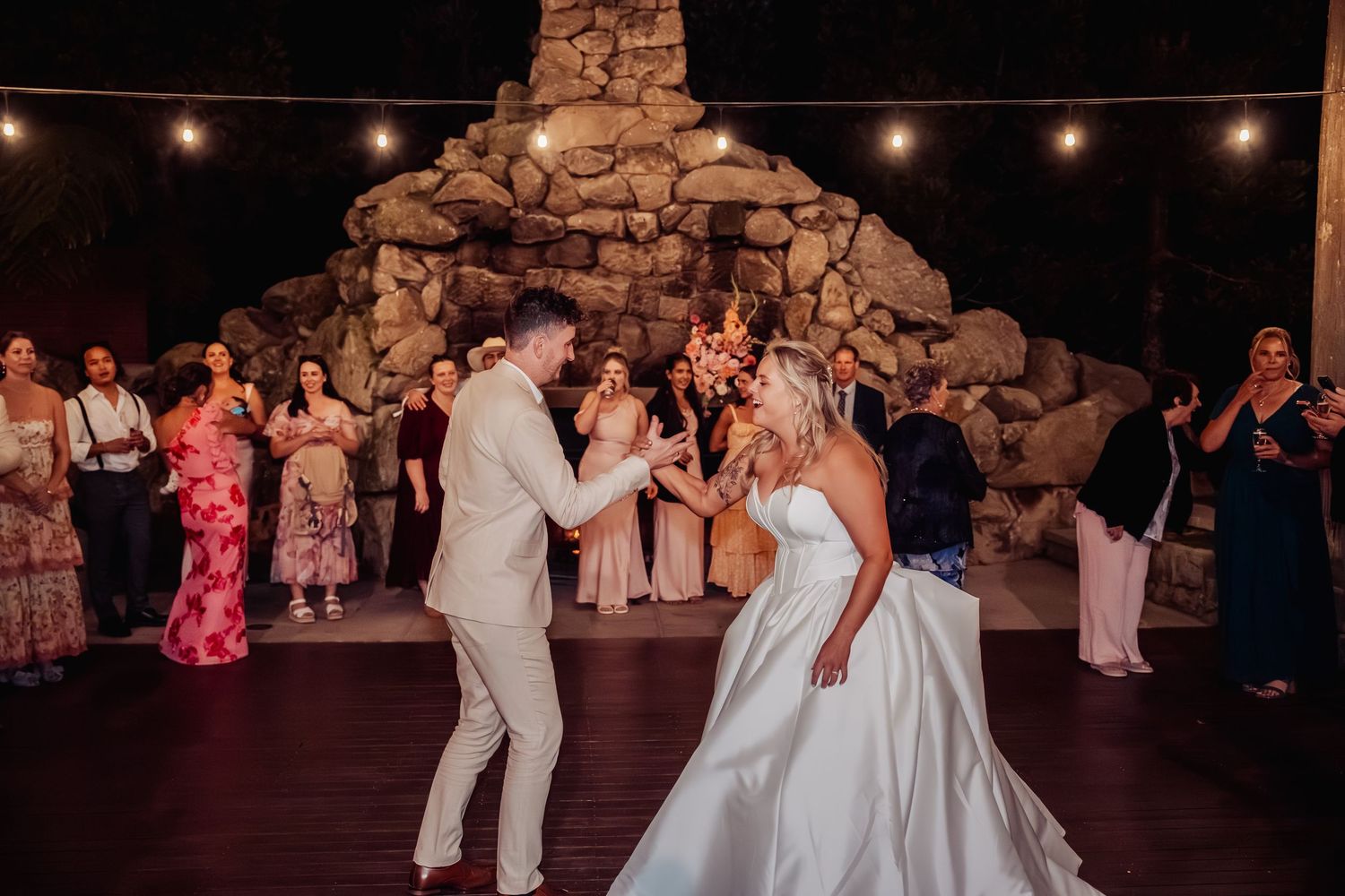 Bride and groom share first dance under string lights before stone fireplace surrounded by wedding guests.