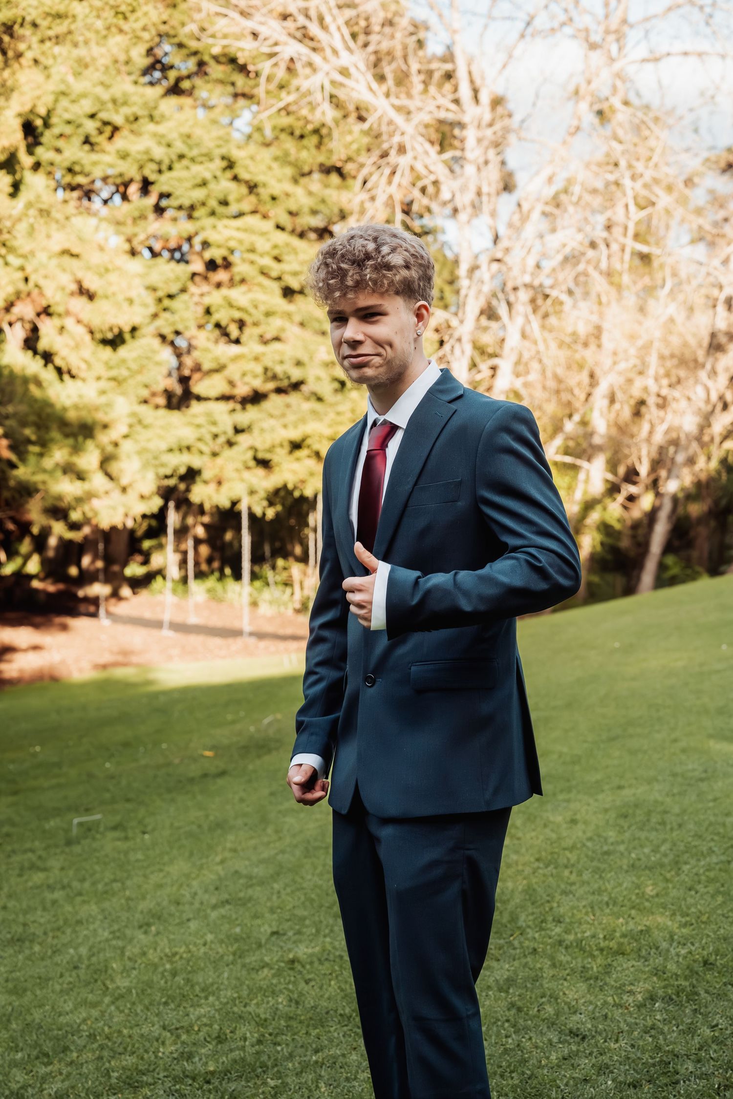 Young groom in navy suit and red tie stands on green lawn with trees in background.