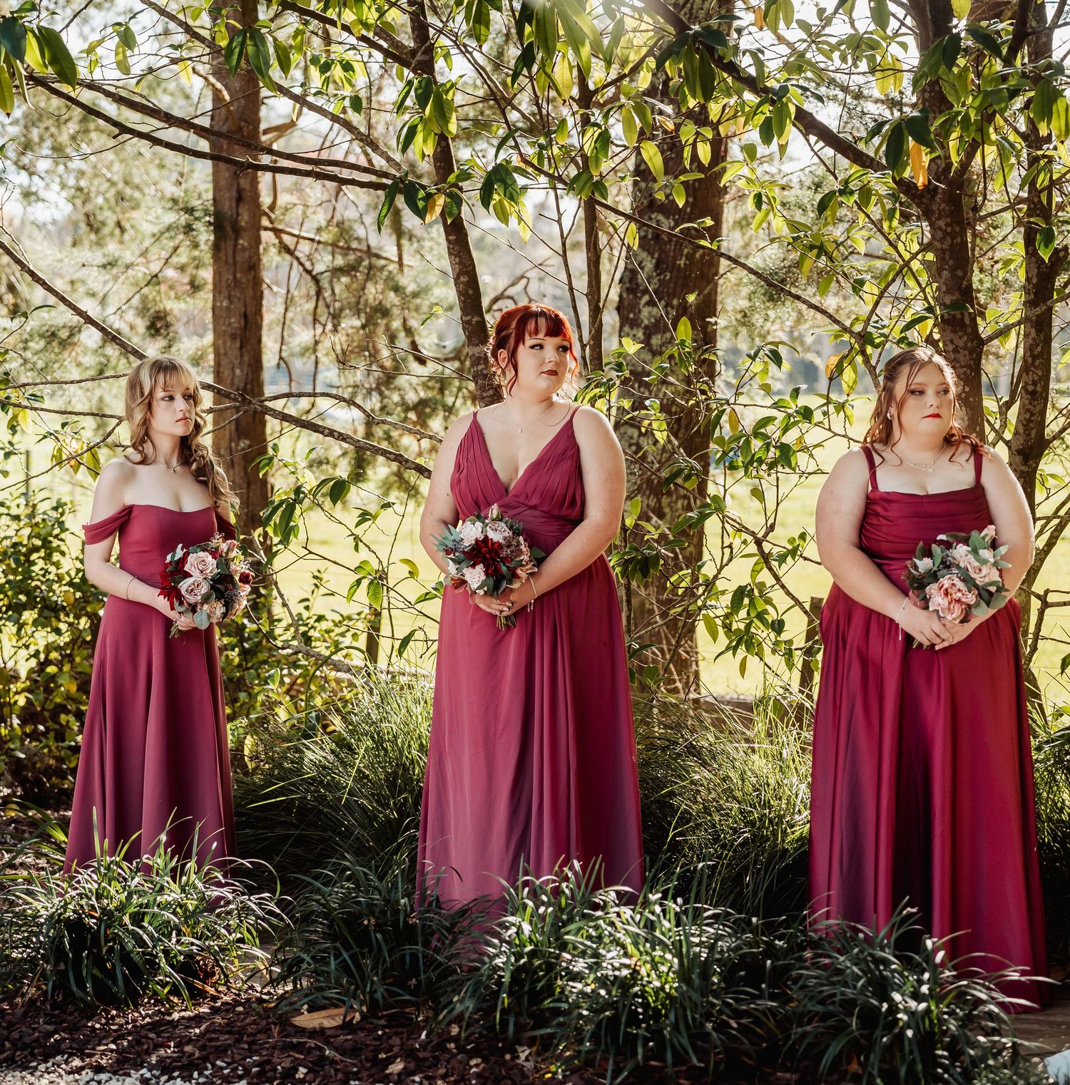 Three bridesmaids in burgundy gowns holding bouquets standing among trees in dappled light.