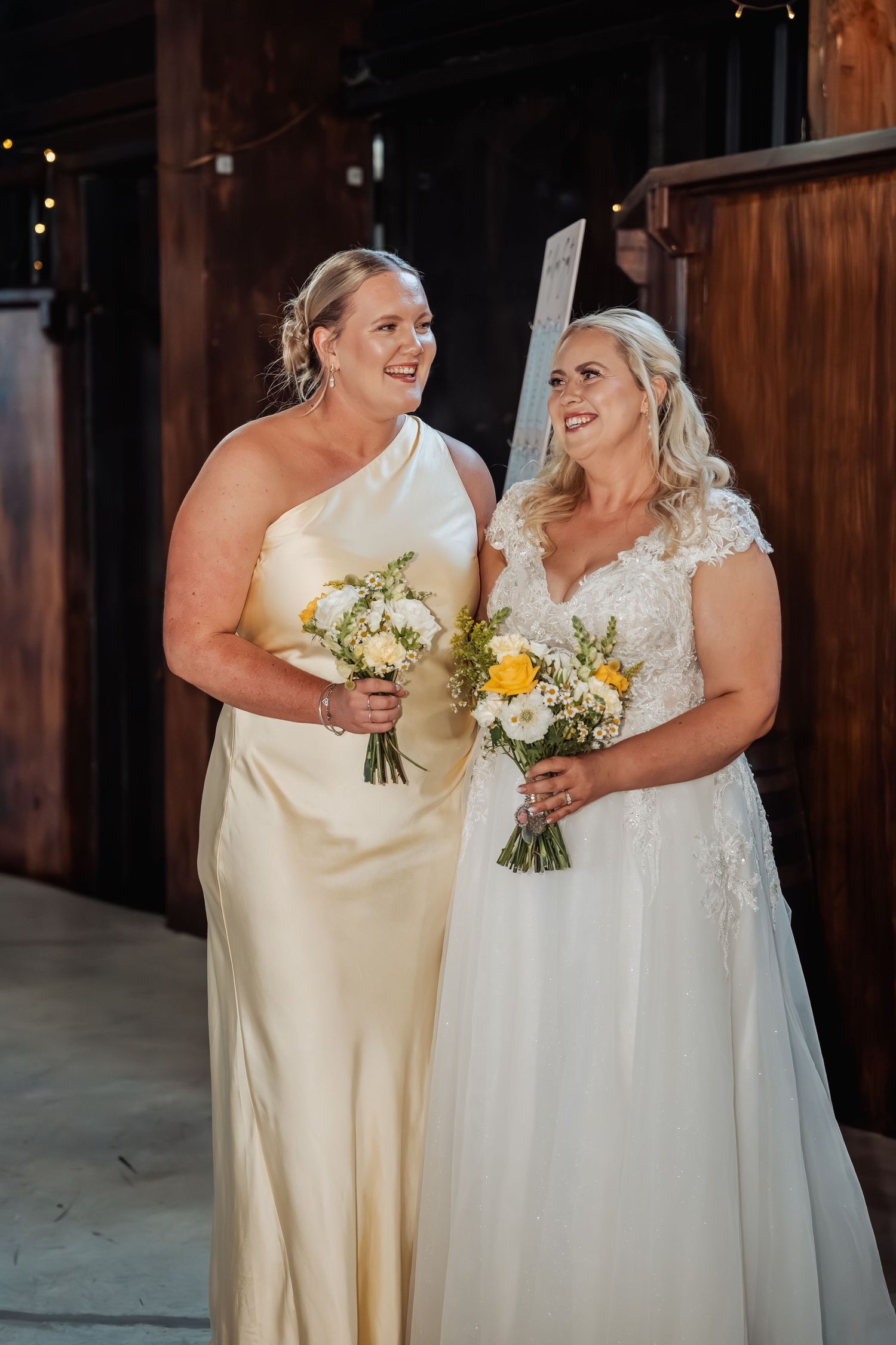 Bride in lace gown and bridesmaid in champagne dress holding yellow bouquets in barn.