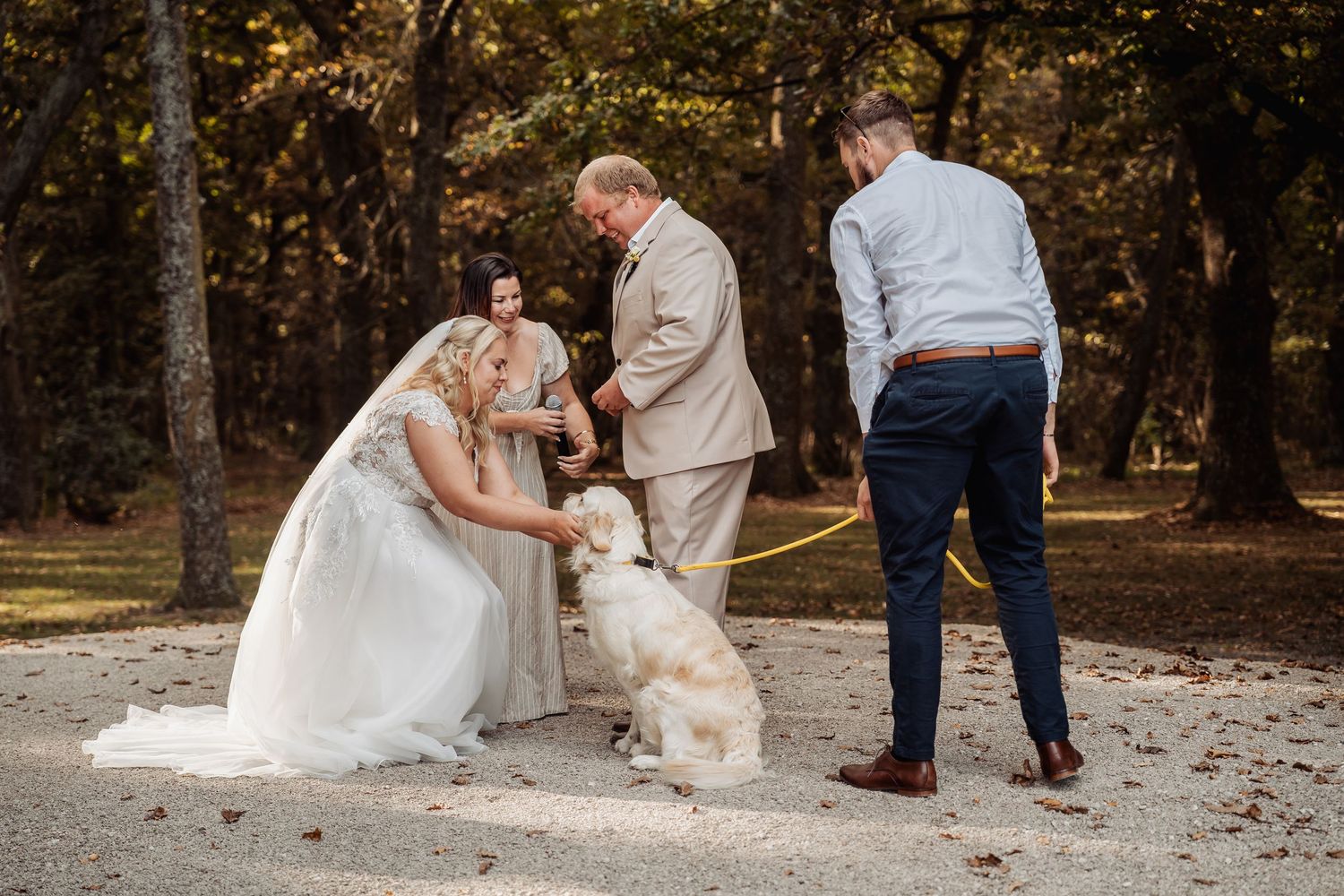 Bride and groom kneeling to greet their white dog during outdoor wedding photo session.