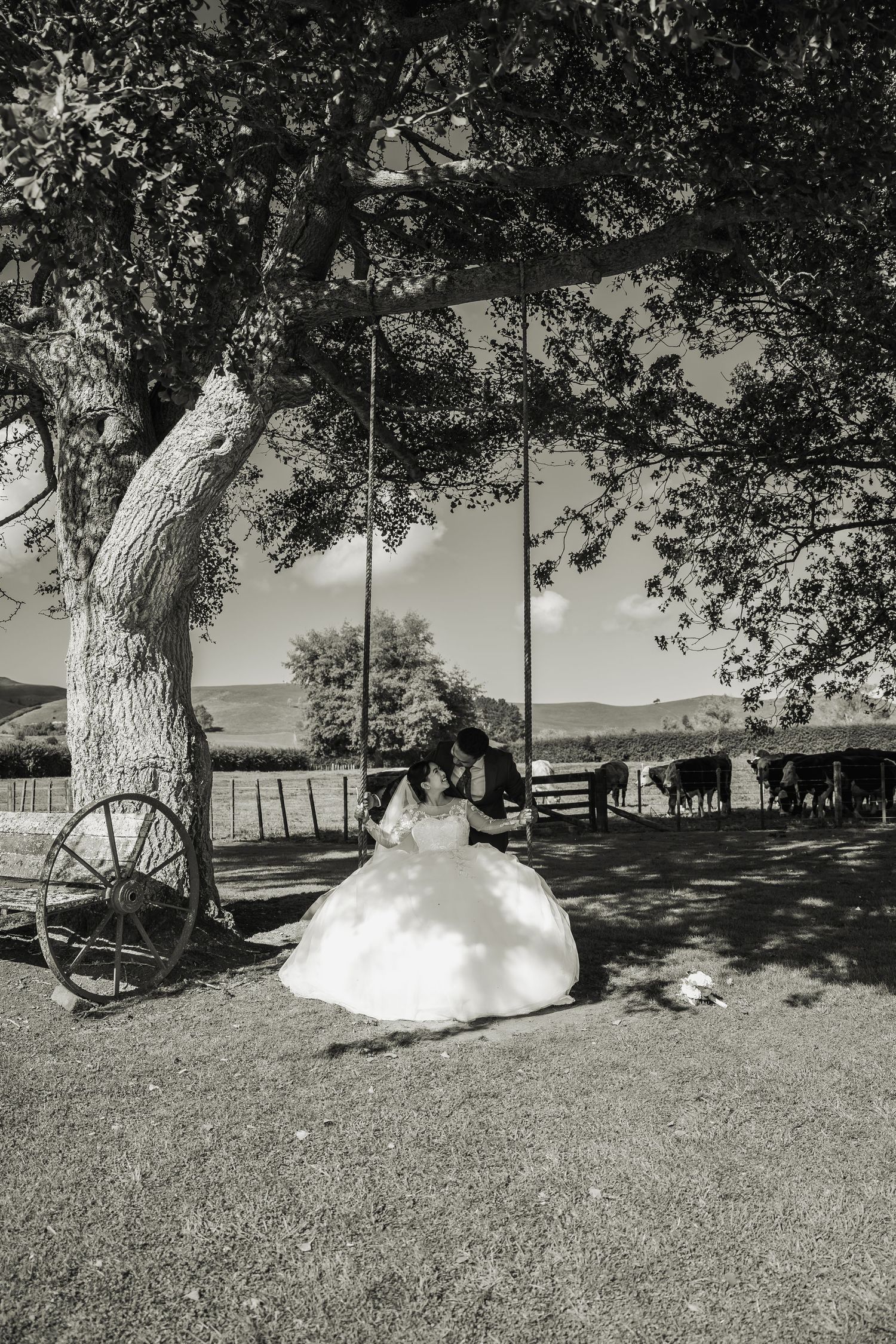 Black and white photo of bride and groom sharing romantic moment on swing under large tree.