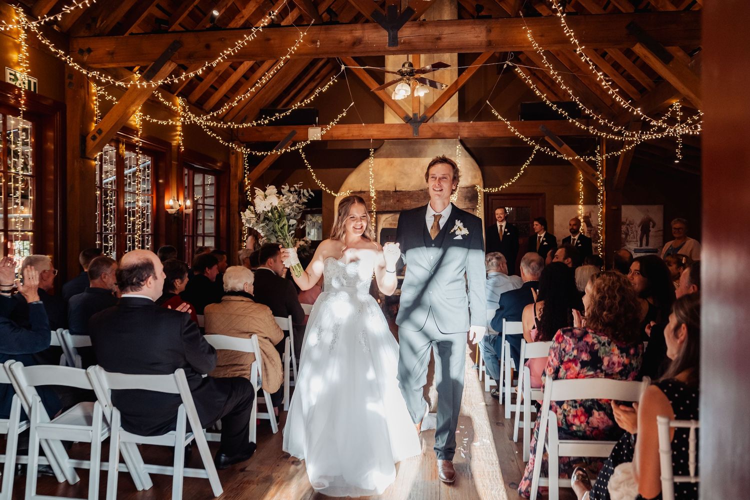A newlywed couple walks down the aisle in a rustic barn venue decorated with twinkling lights and white chairs.