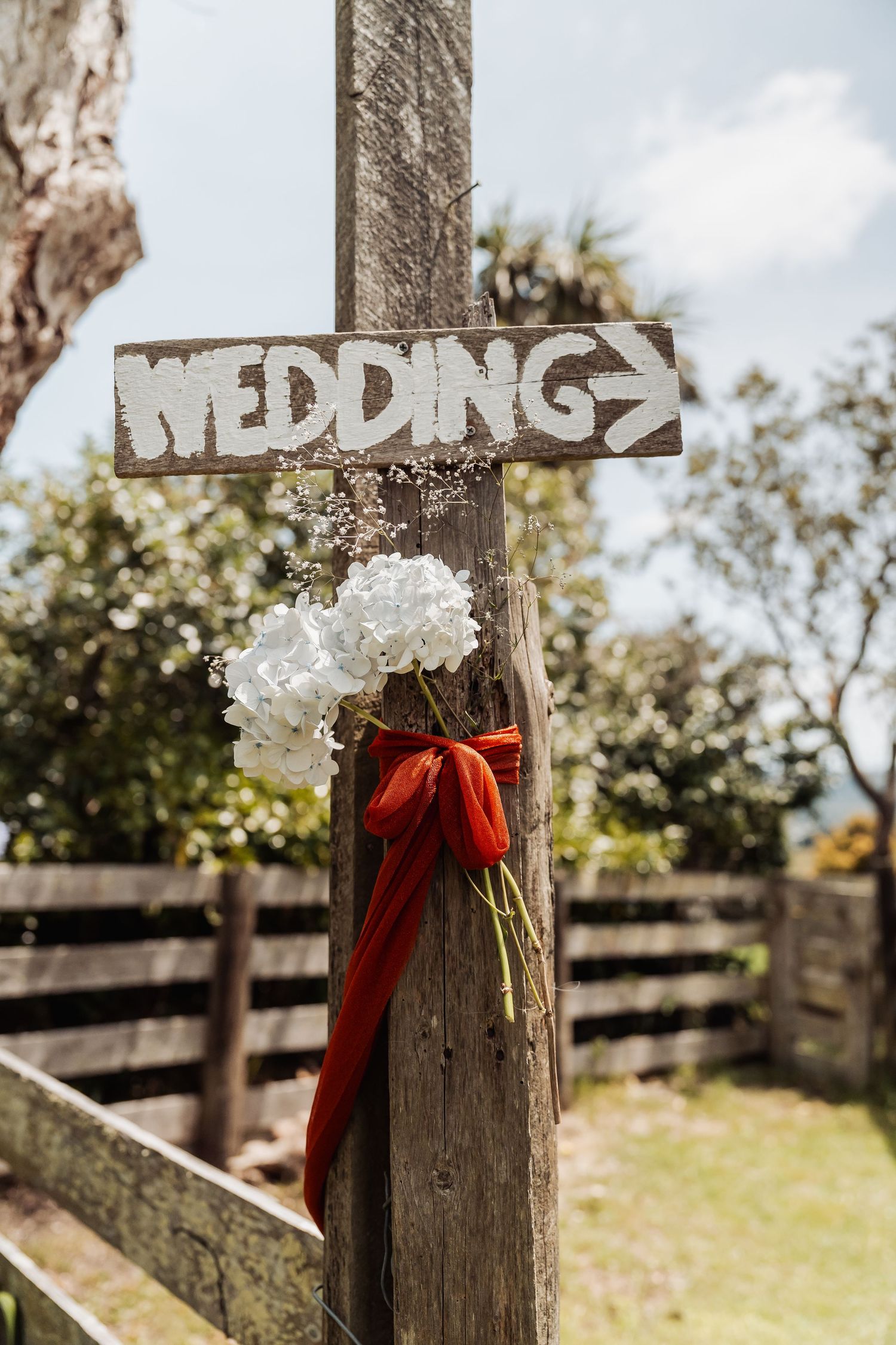 Rustic wooden wedding sign with white hydrangeas and red tulips tied with orange ribbon on a fence post.