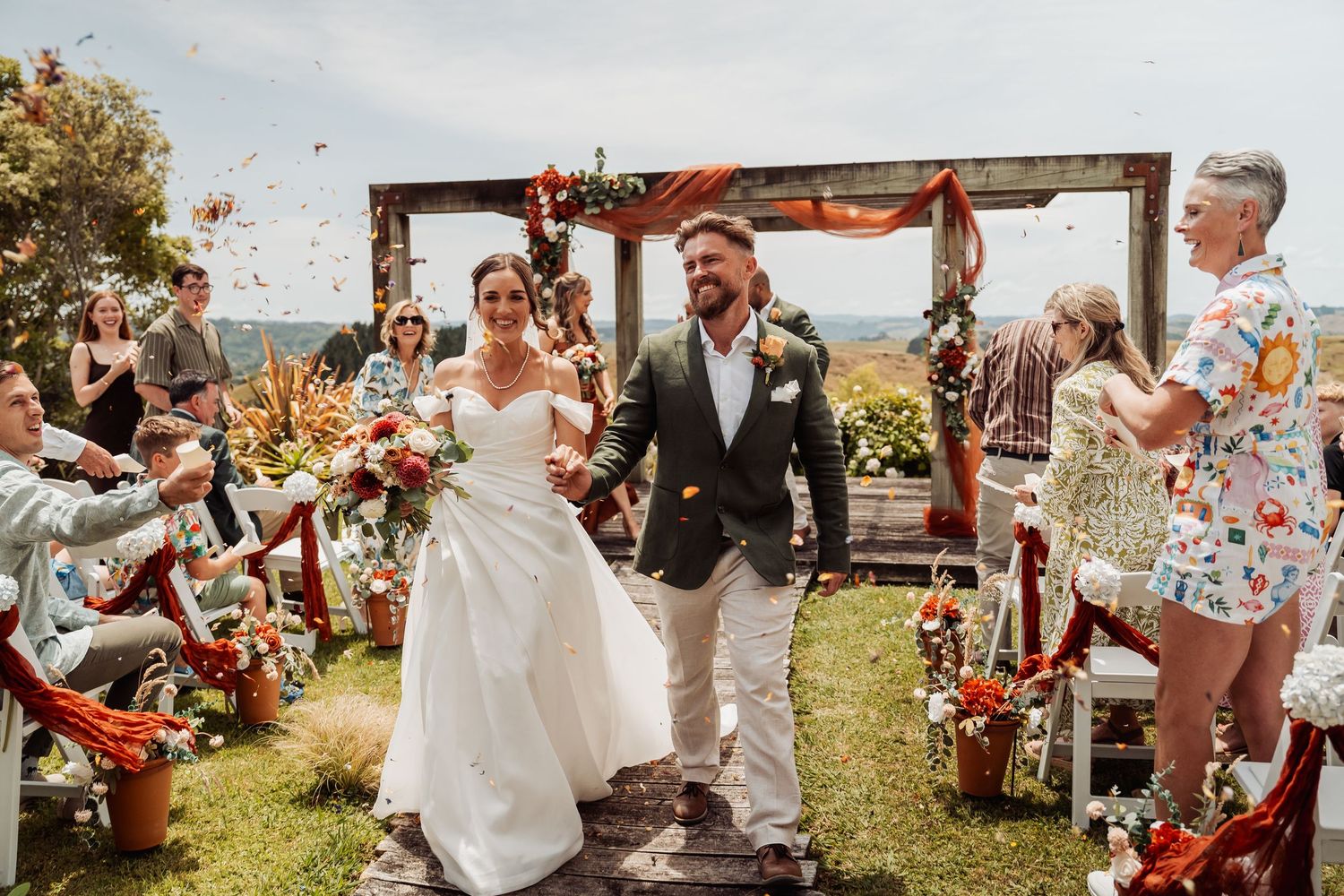 Bride and groom walk down outdoor aisle as guests toss confetti at a vibrant tropical wedding ceremony.