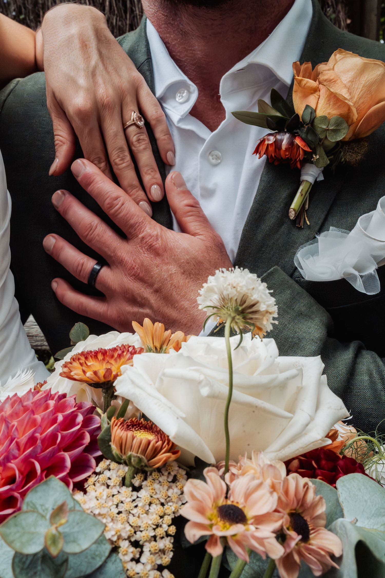 Newlyweds displaying wedding rings over a vibrant bridal bouquet of roses and dahlias.