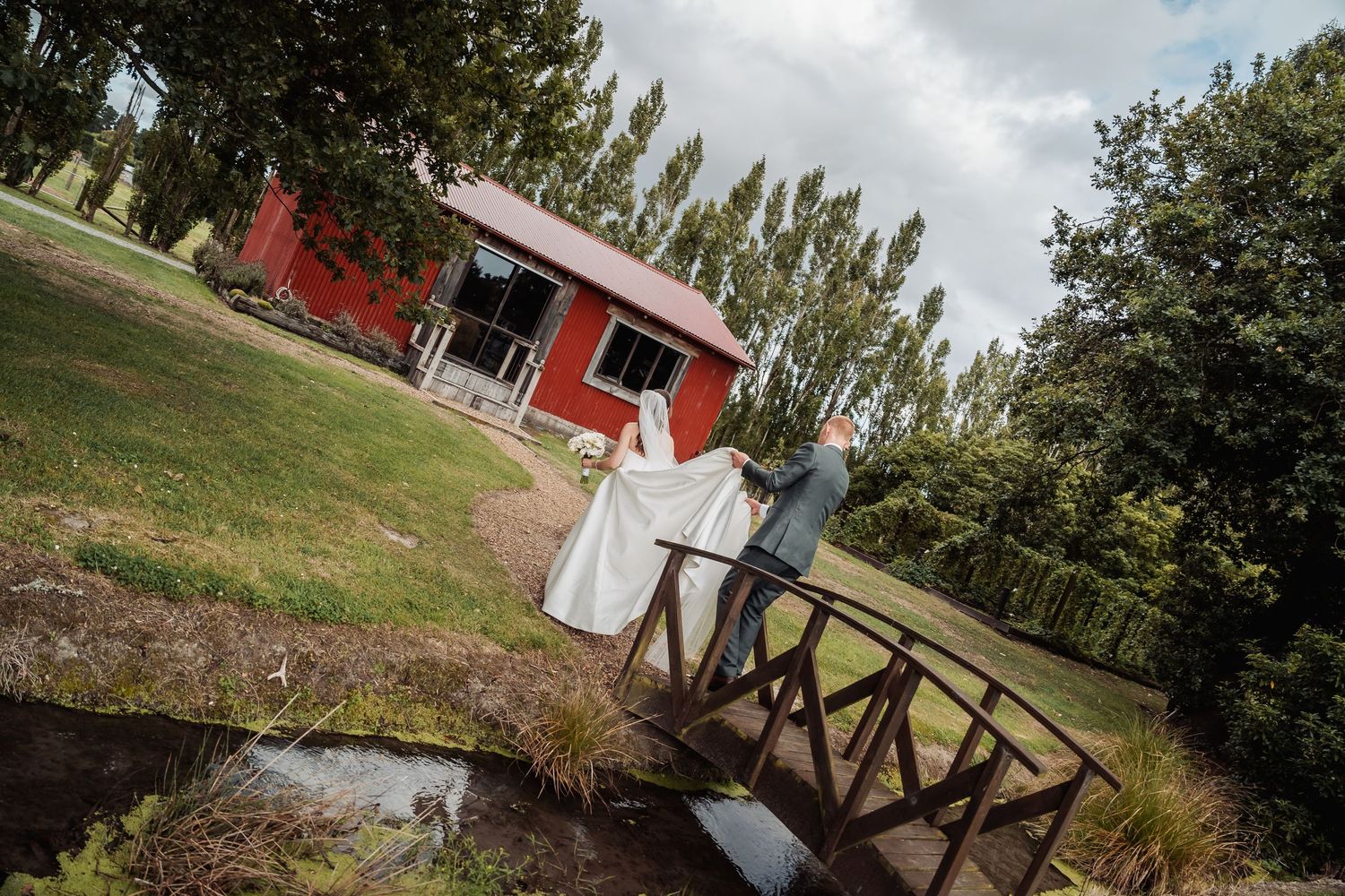 Bride and groom cross a wooden bridge near a red barn surrounded by lush green trees and gardens.