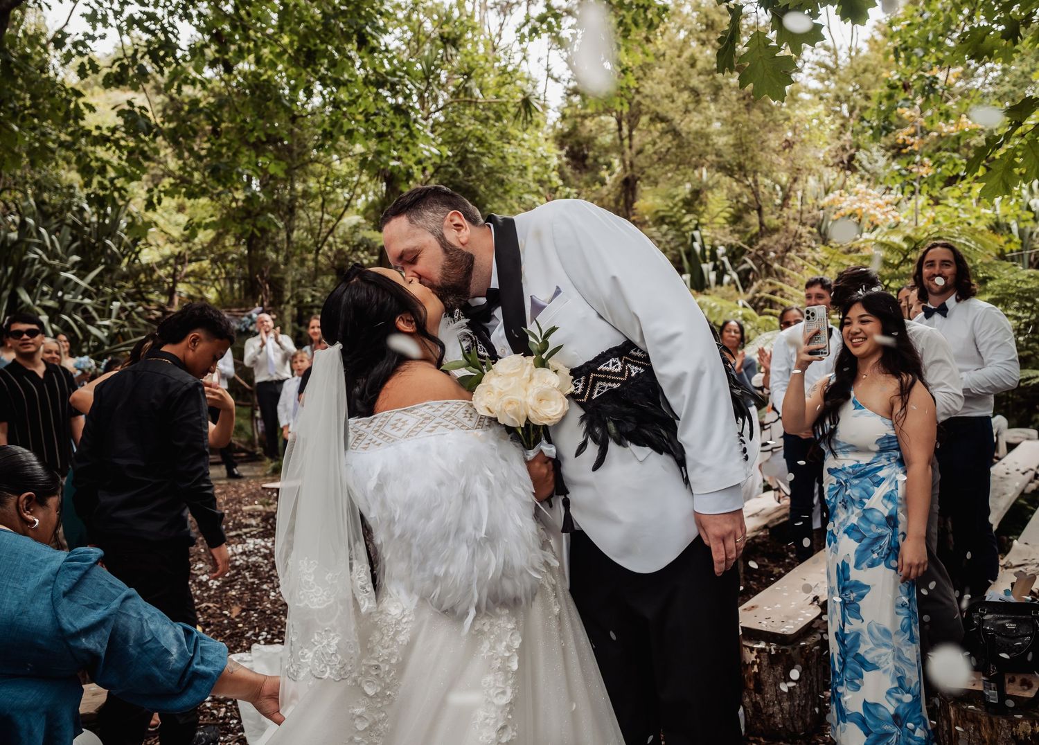 Newlyweds share first kiss surrounded by cheering guests at lush green forest wedding ceremony.