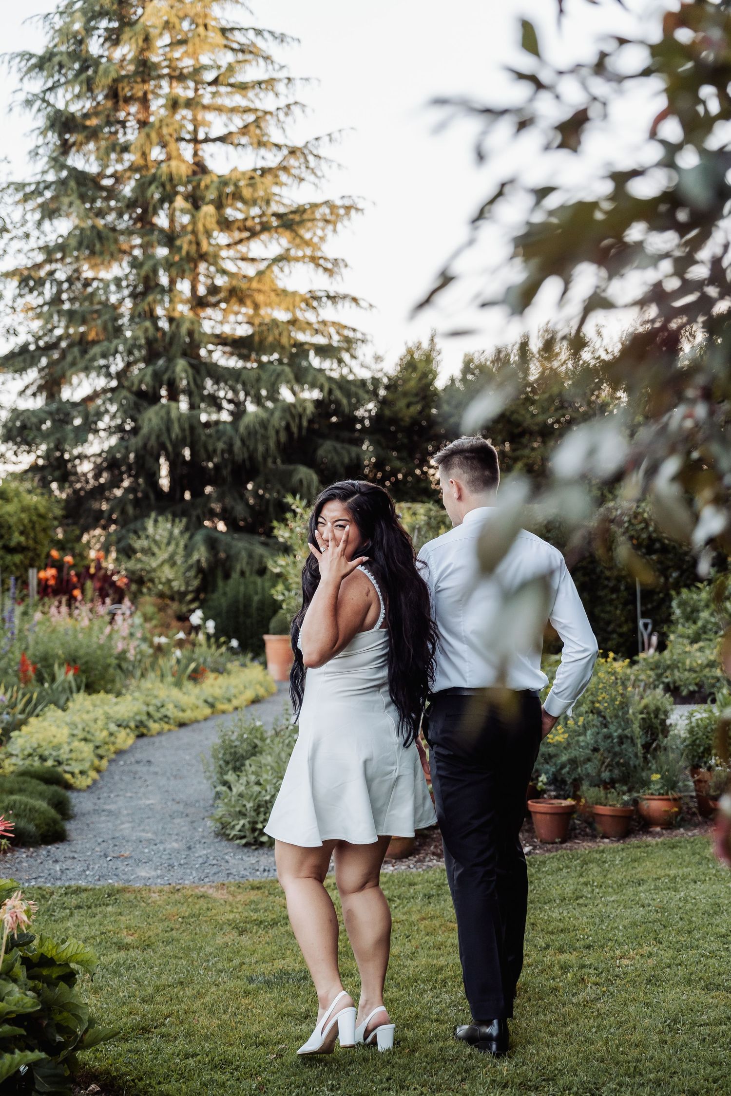 Bride in white mini dress laughs while walking with groom through lush garden path at sunset.