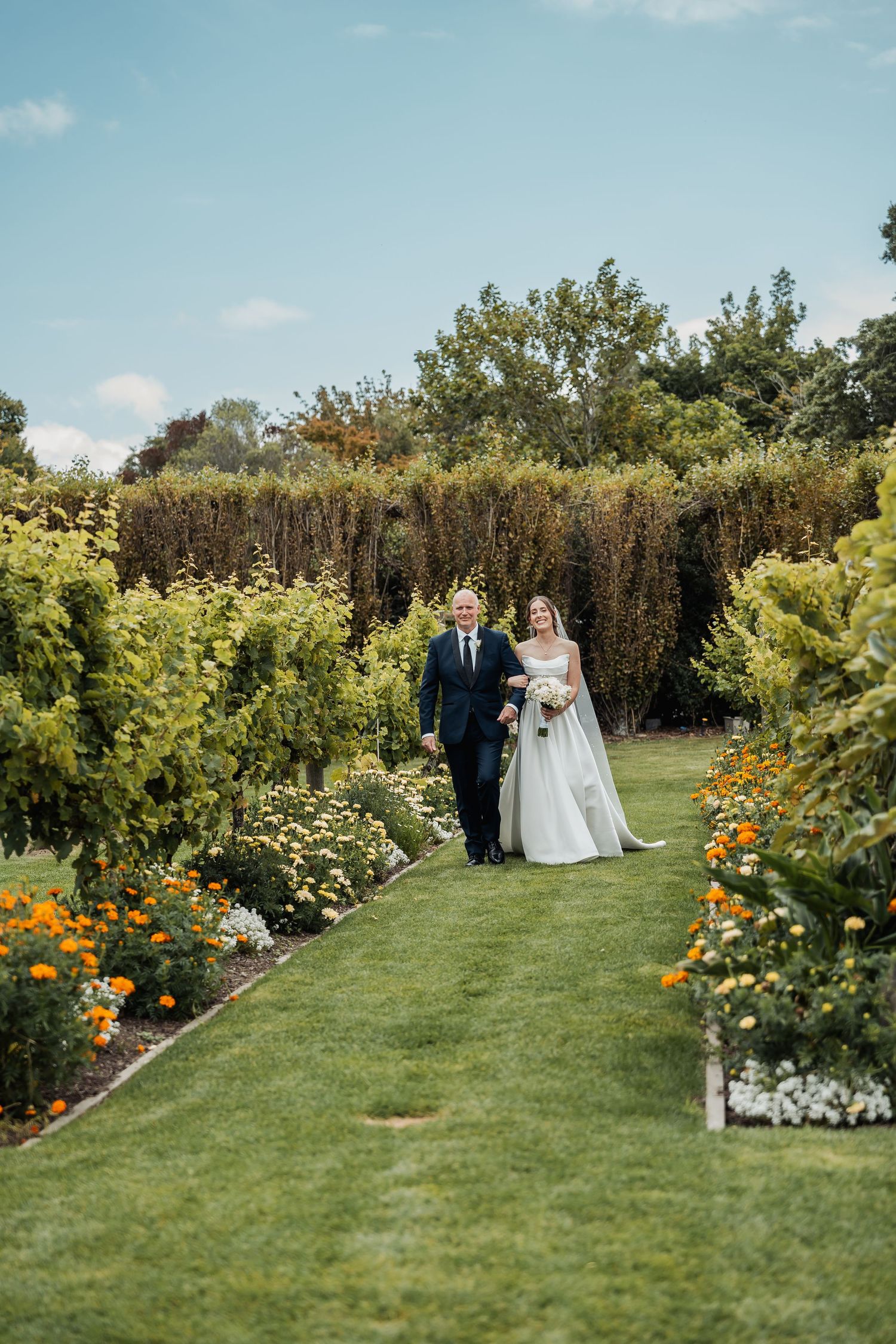 Bride and her father walk down a lush garden aisle lined with colorful flowers and greenery.