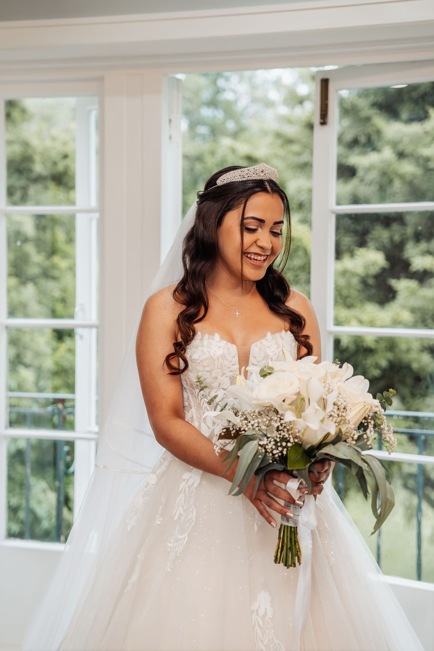 Radiant bride in lace ballgown holding white floral bouquet near bright windows.