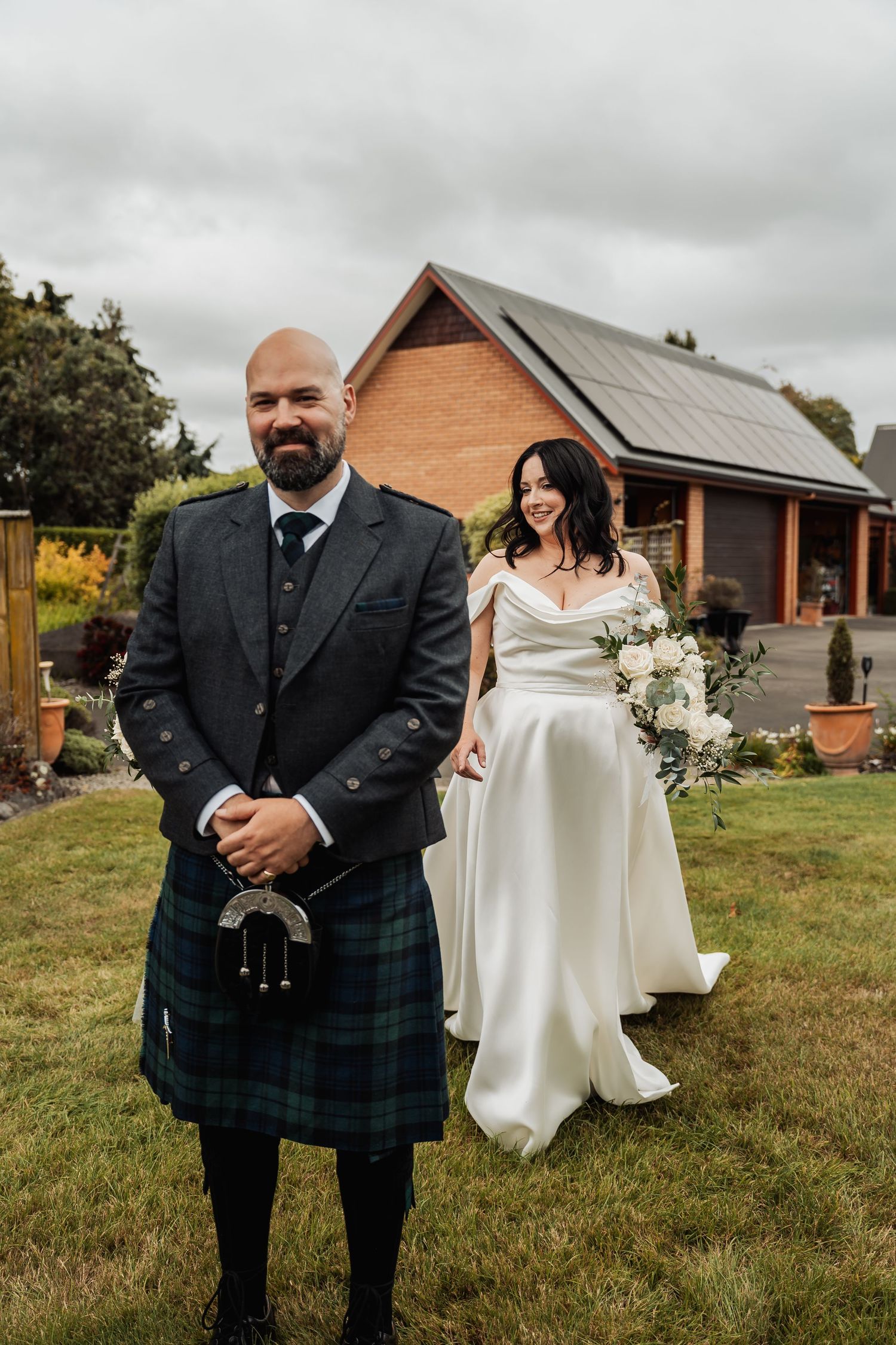 A bride in a white off-shoulder gown holds a bouquet while standing behind a groom in a Scottish kilt outdoors.