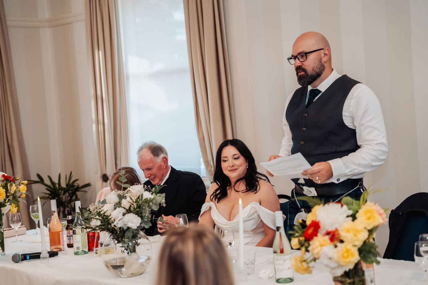 Bald groom in vest reads speech while smiling bride sits at elegantly decorated wedding reception table.