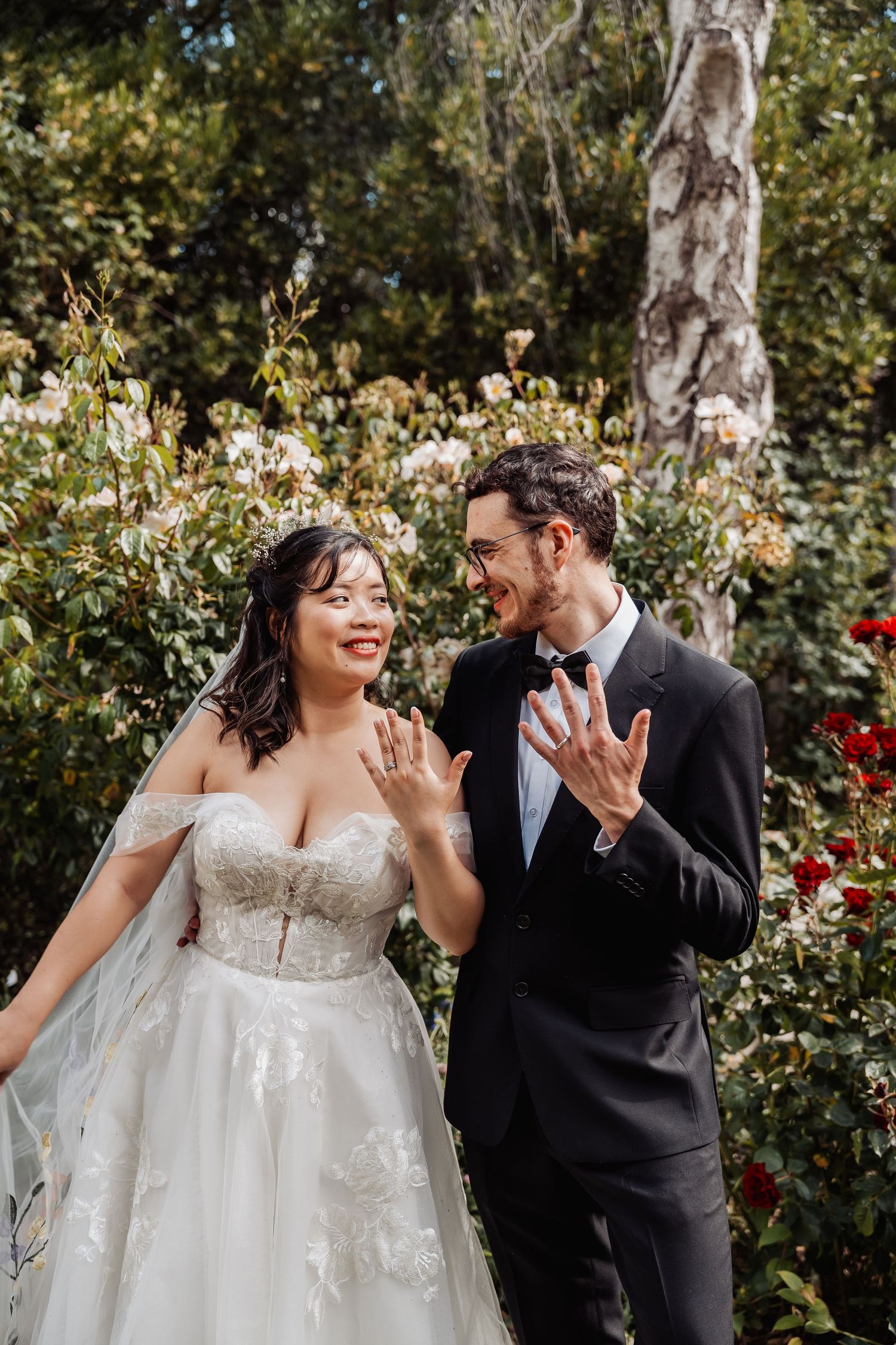 Newlyweds in garden showing wedding rings, bride in lace gown with veil, groom in black suit, surrounded by roses.