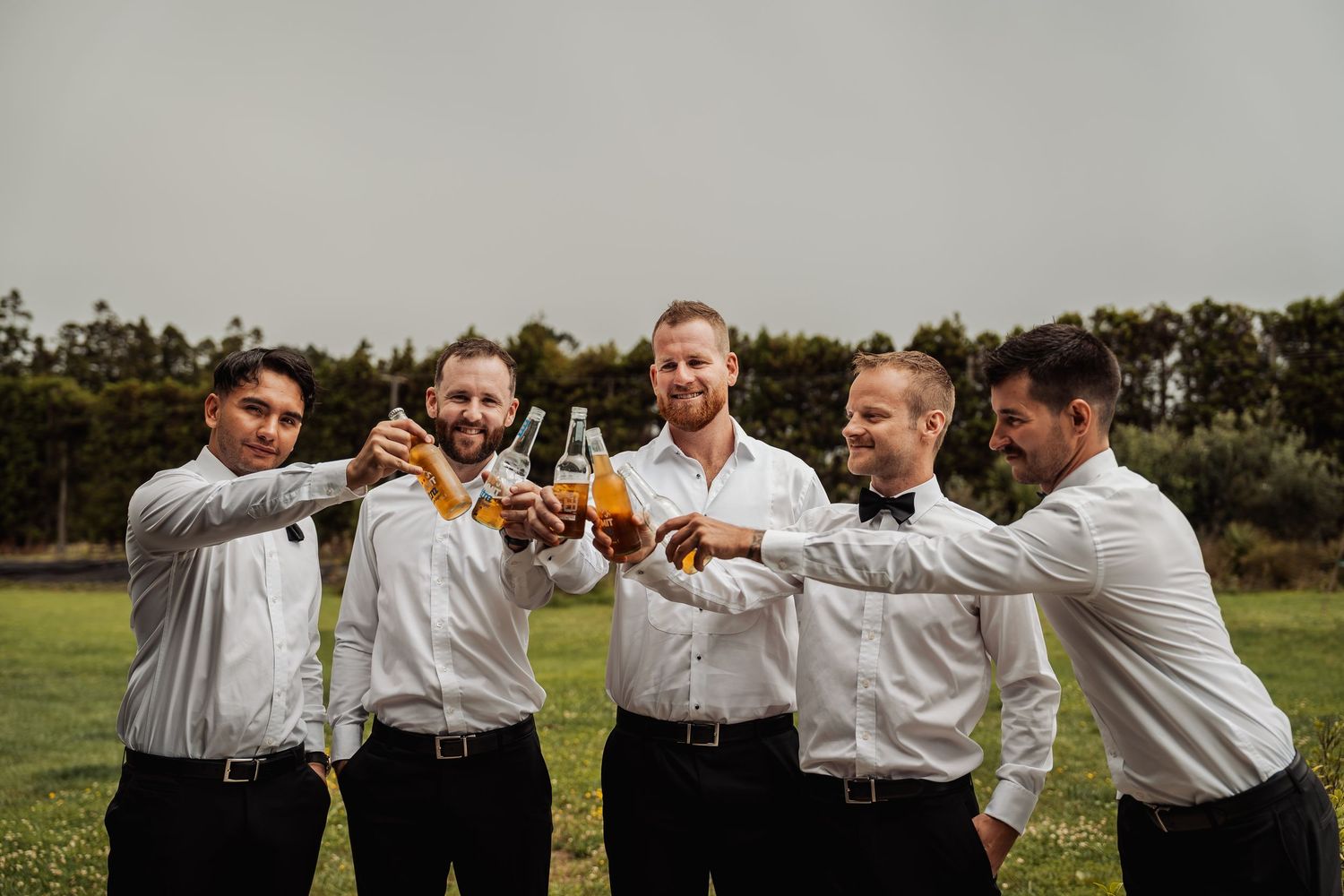 Five groomsmen in white shirts and black pants toasting with beer bottles on a green lawn.