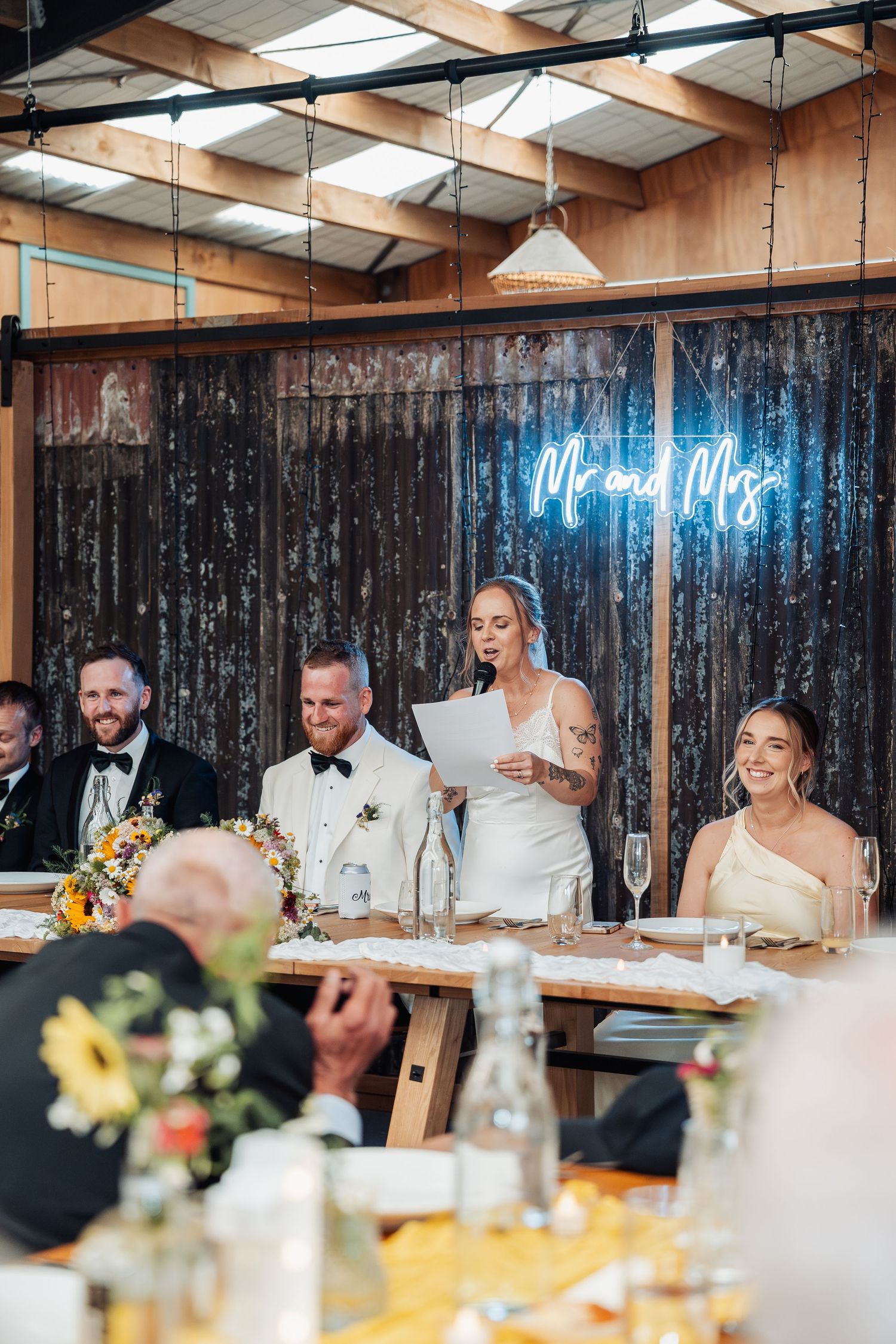 Bride giving a speech at rustic barn wedding reception with neon Mr and Mrs sign, groom and bridal party seated at table.