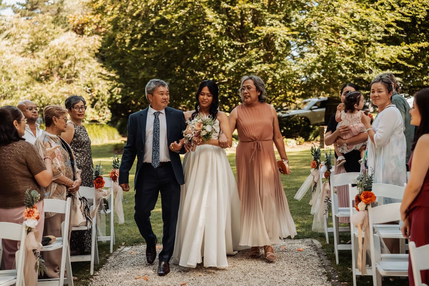 Bride walks down outdoor aisle escorted by two family members past seated wedding guests.