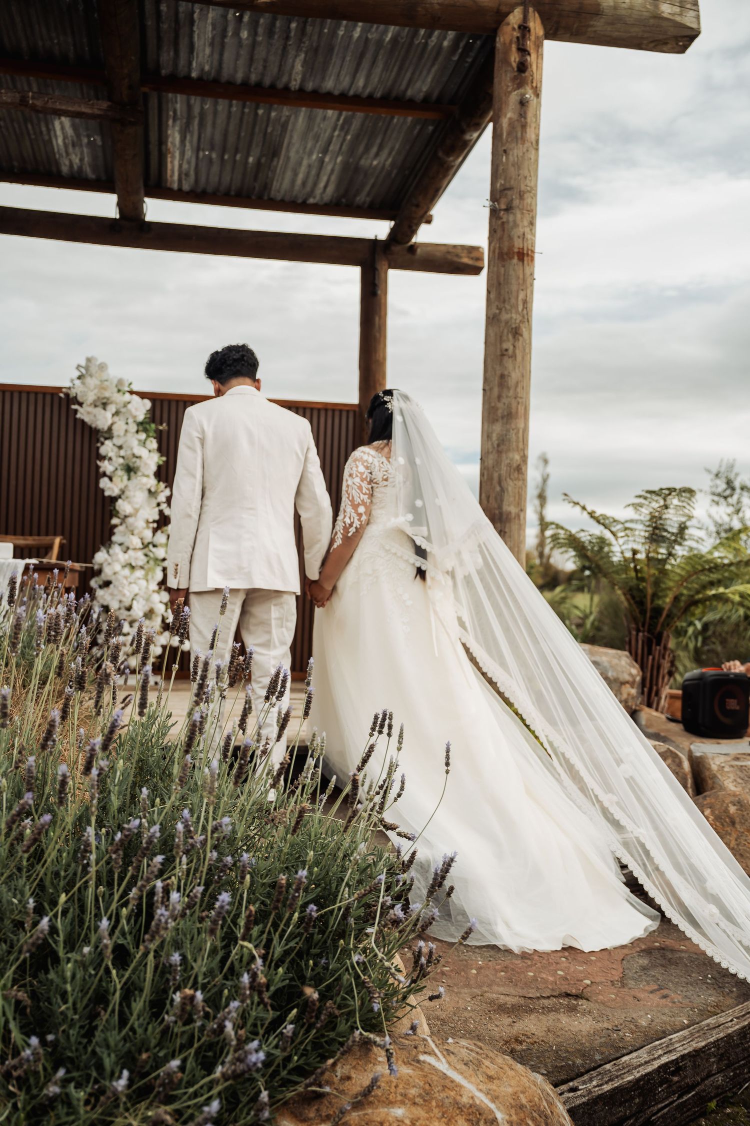 Bride and groom walking hand in hand under a rustic wooden pergola surrounded by lavender and white floral arrangements.
